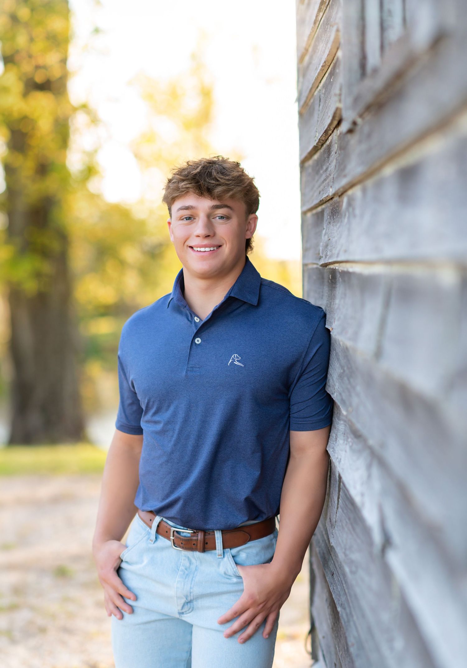 Smiling teenage boy in blue polo shirt and jeans leaning against rustic wooden wall outdoors.