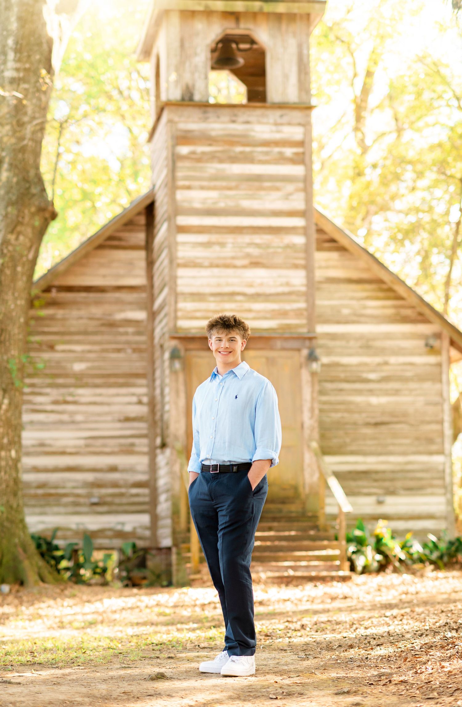 Teen boy in light blue shirt and navy pants smiling in front of rustic wooden chapel outdoors.