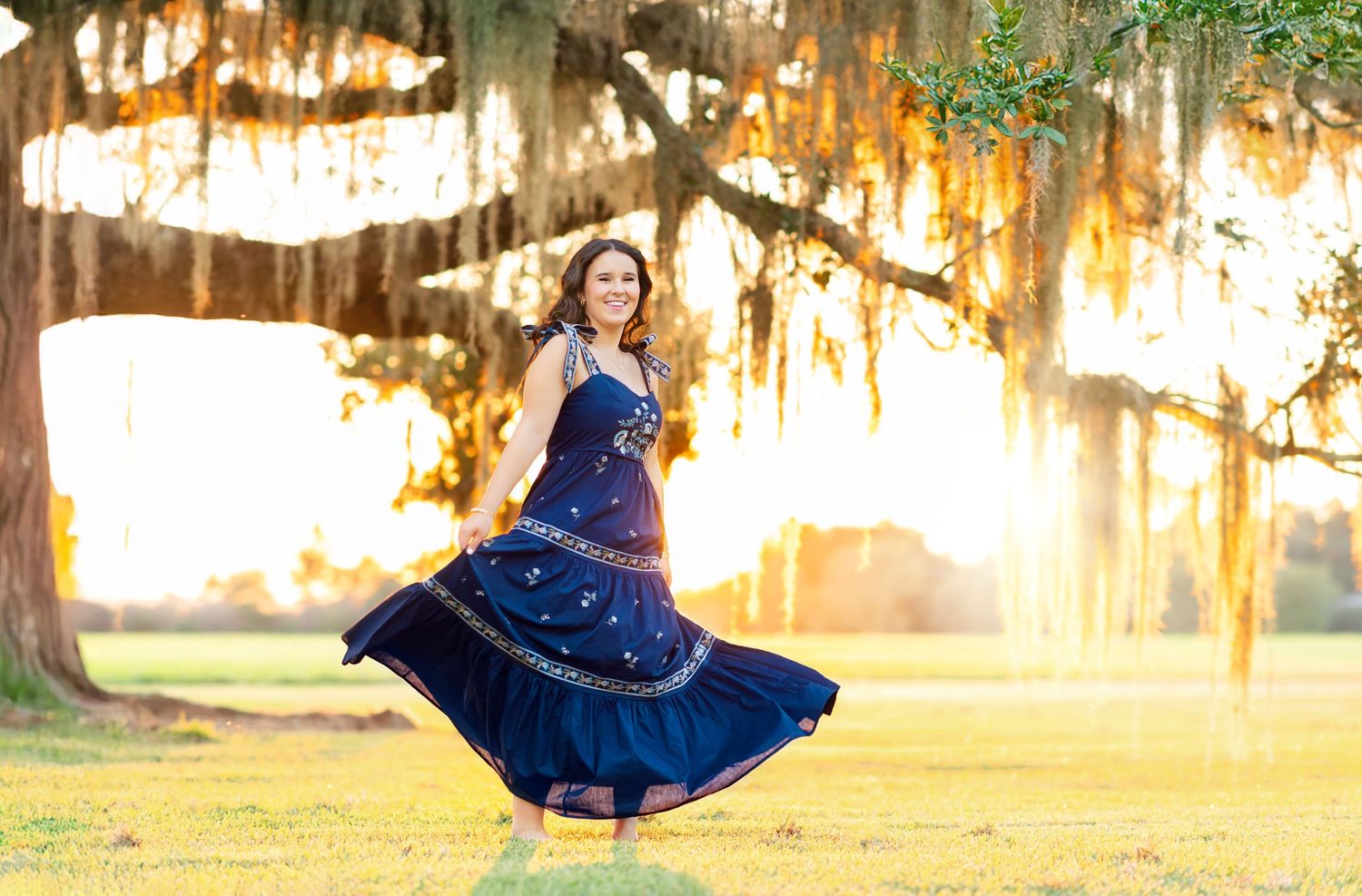 Woman in navy blue gown twirling under Spanish moss-draped oak tree at golden sunset in a lush green park.