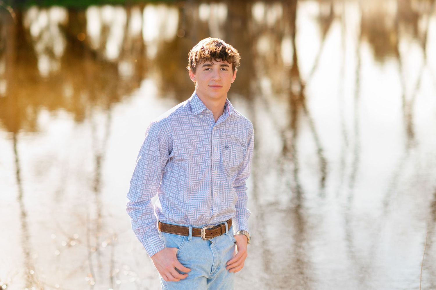 Young man in light blue shirt and jeans poses outdoors near reflective water during golden hour.