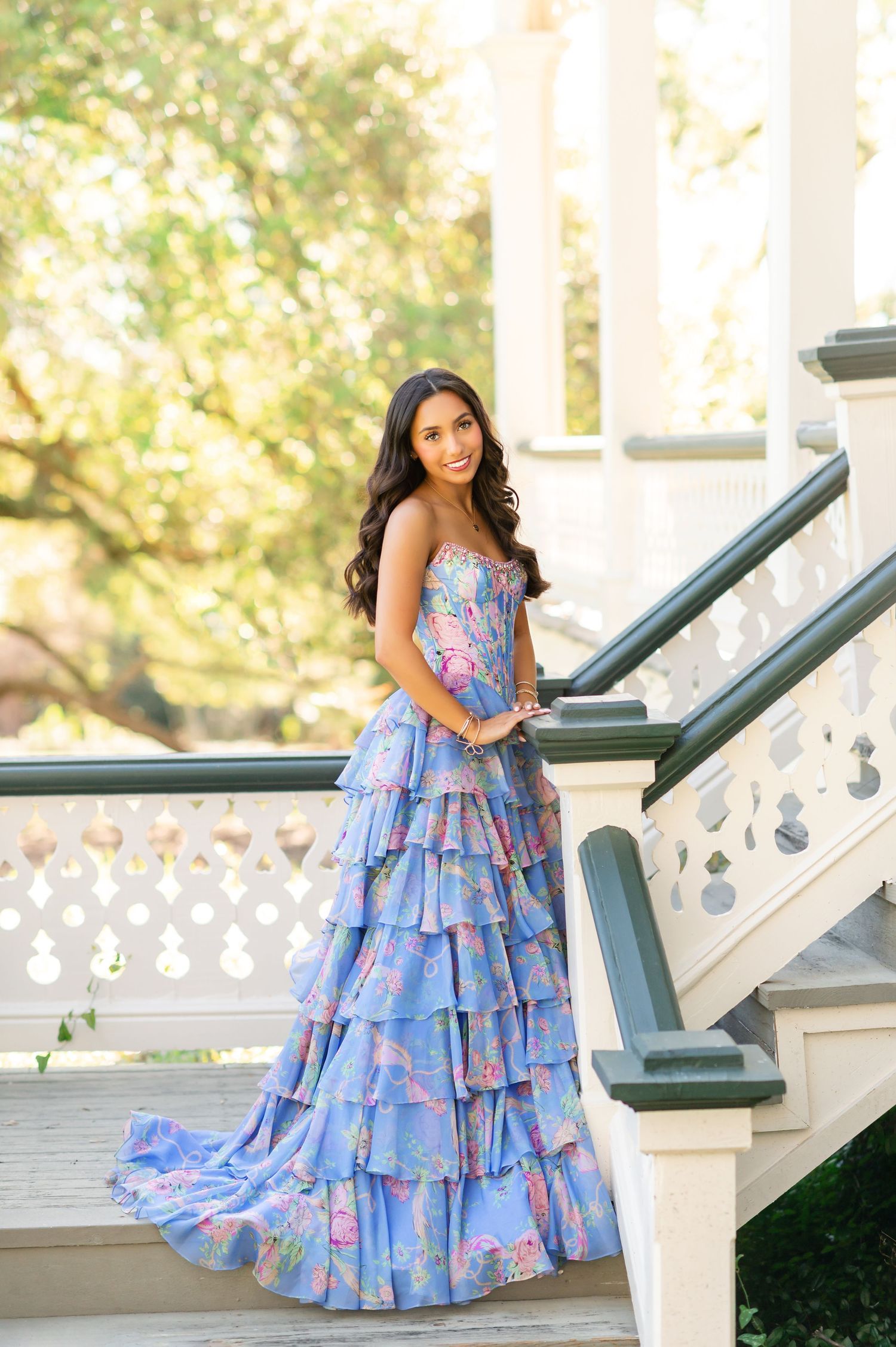 Young woman in a blue floral tiered ruffle gown poses gracefully on elegant white staircase outdoors.