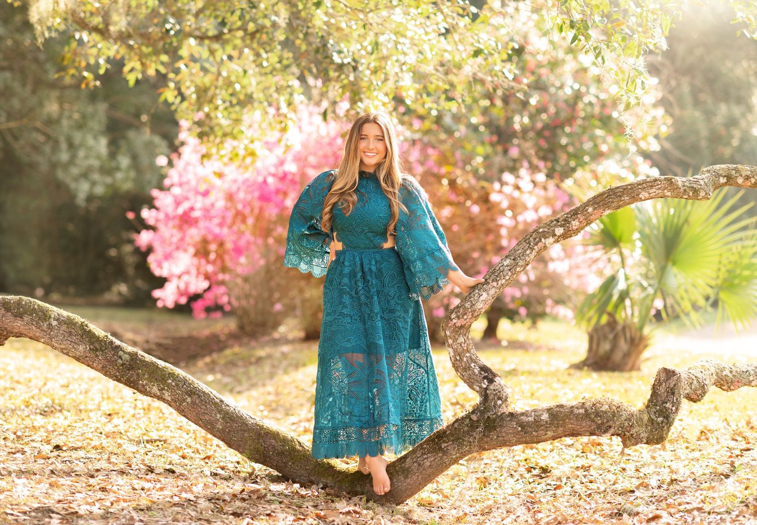 Woman in teal lace dress standing on tree branch surrounded by pink blooming flowers in a sunlit garden.