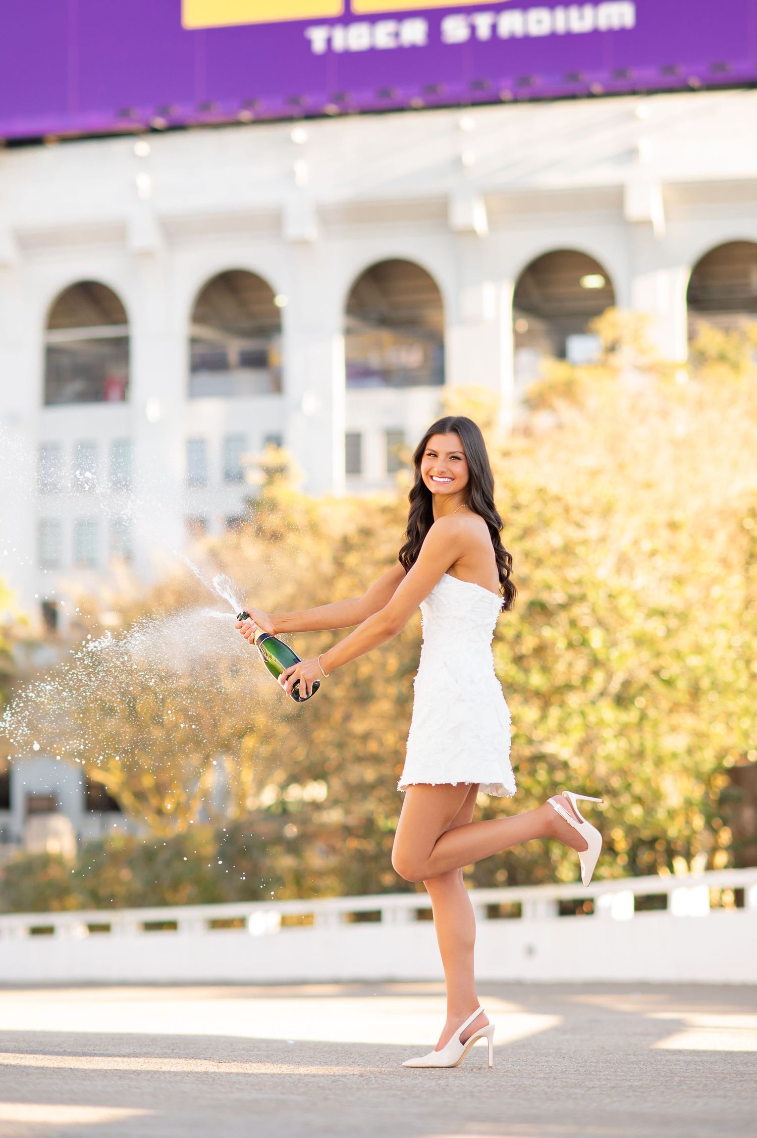 Woman in white dress popping champagne at LSU Tiger Stadium.