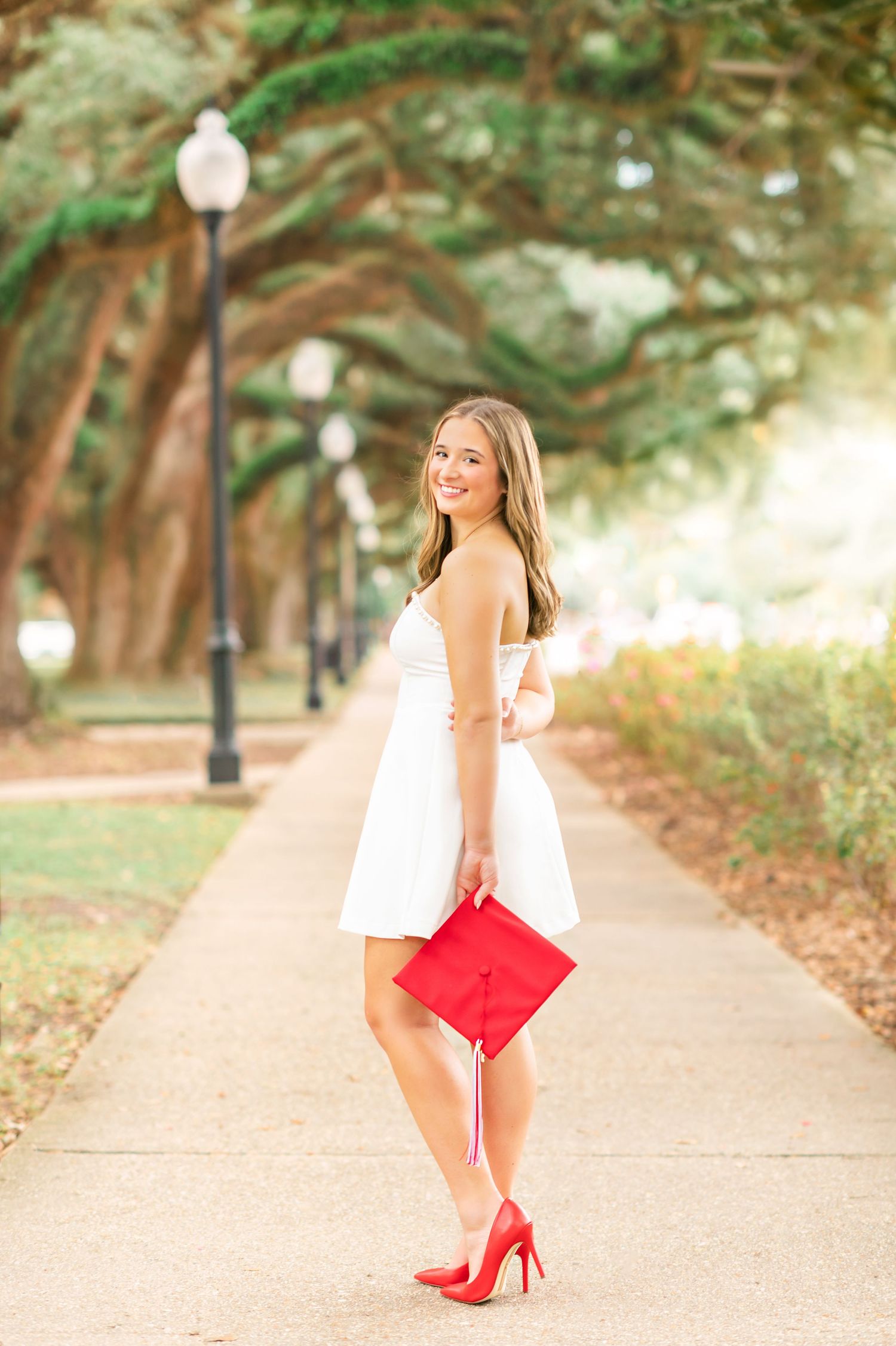 Smiling graduate in white dress and red heels holds red graduation cap on a tree-lined campus walkway.