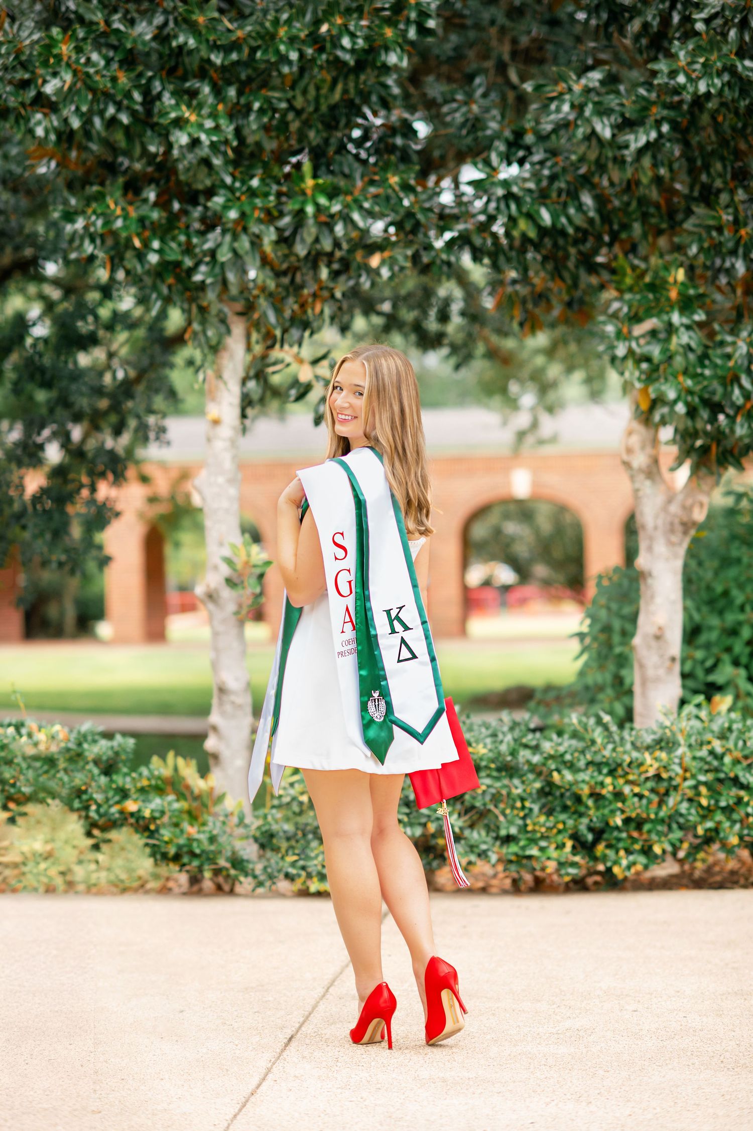 Smiling blonde graduate wearing KD sorority and SGA sashes in white dress with red heels outdoors on campus.