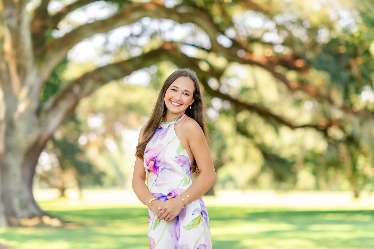 Happy young woman in colorful floral dress standing beneath sprawling oak tree outdoors.