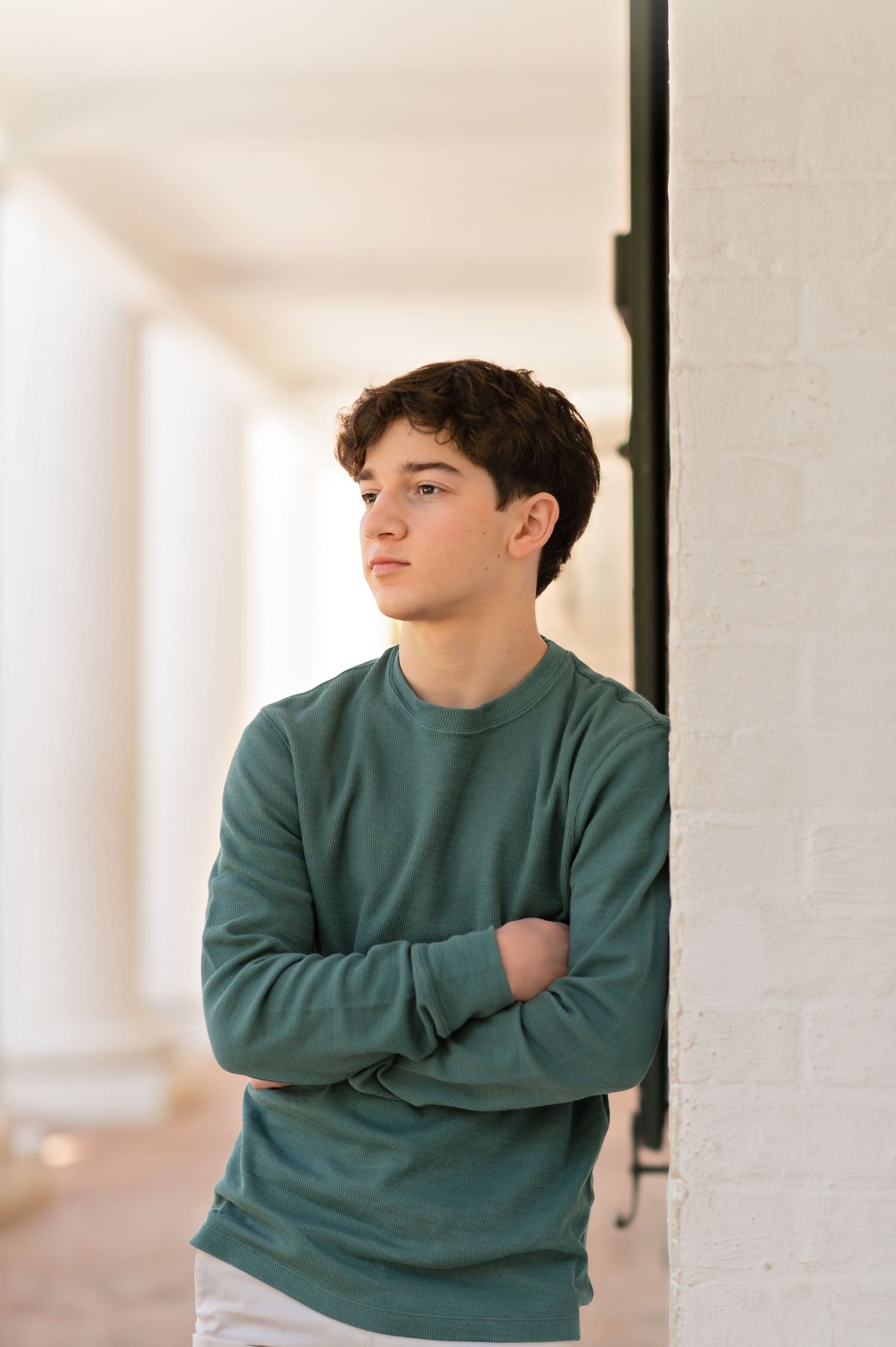 Teen boy in green sweatshirt leans against white brick wall with arms crossed, gazing thoughtfully down a bright hallway.