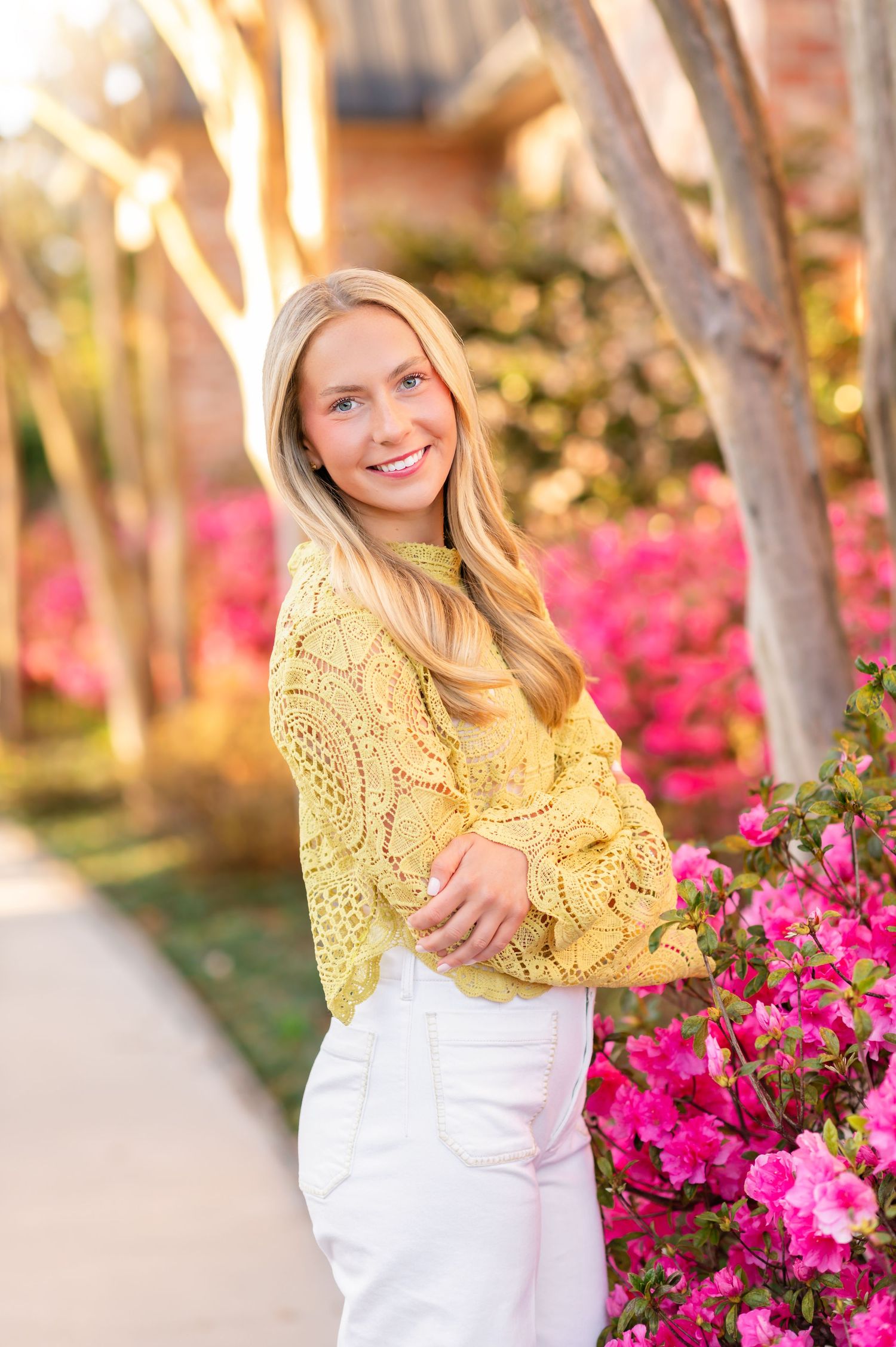 Smiling blonde woman in yellow lace top and white pants posing near pink azalea bushes outdoors.