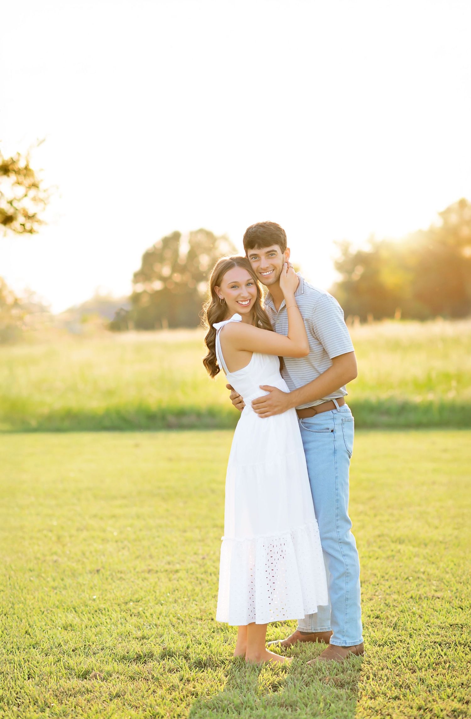 Young couple hugging outdoors at sunset, woman in white midi dress smiling, warm golden light in open field.