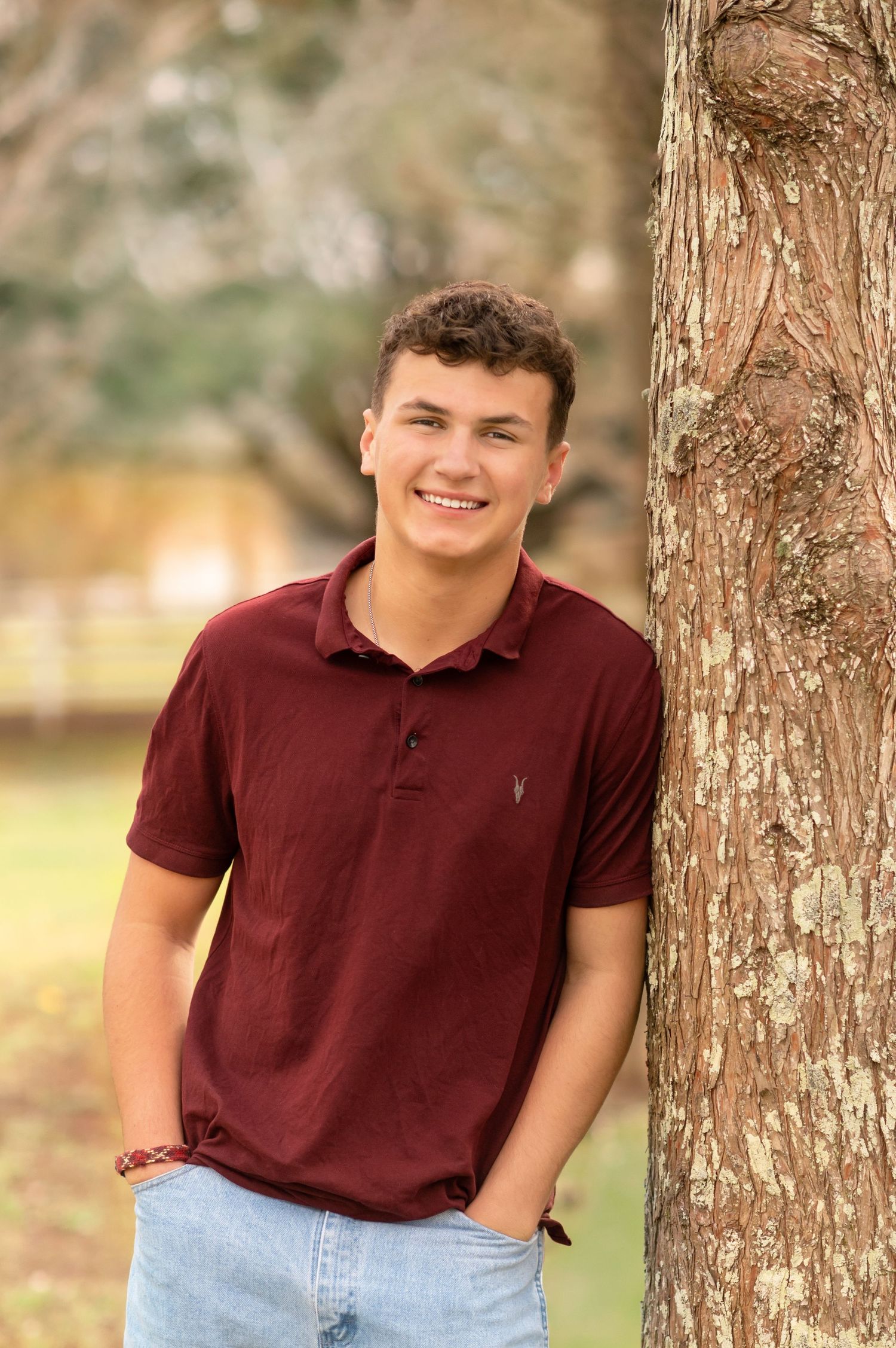 Smiling teenage boy in maroon polo shirt leaning against a tree outdoors for senior portrait photo.