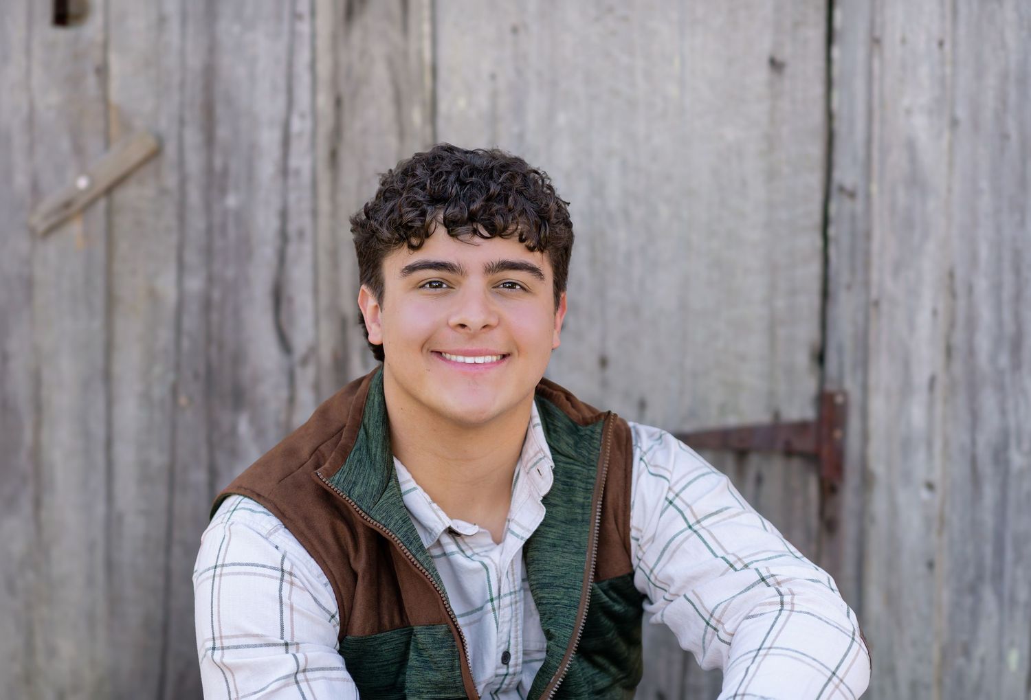 Smiling teenage boy in plaid shirt and brown vest posing outdoors against rustic wooden barn wall.