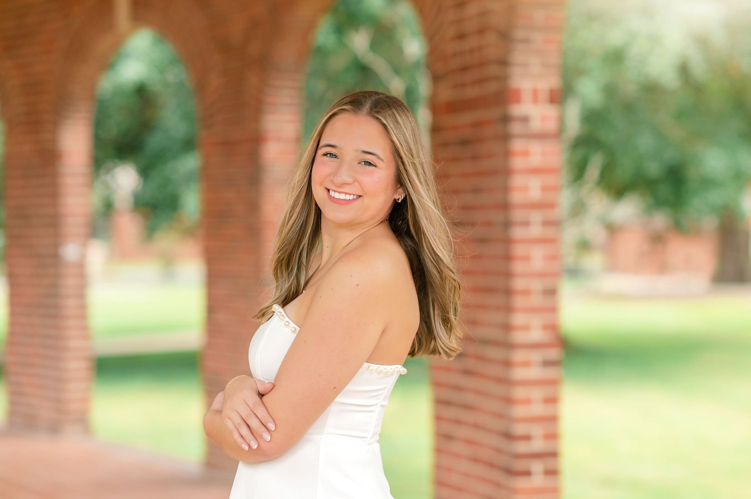 Smiling young woman in white strapless dress posing between brick arched columns outdoors.