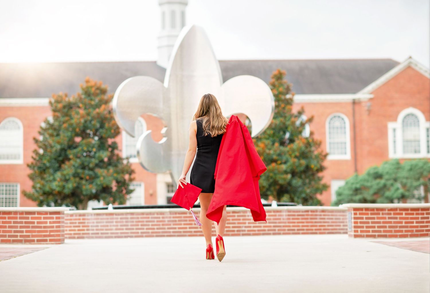 Woman in black dress holding red graduation gown and balloons walks on brick plaza near university building.