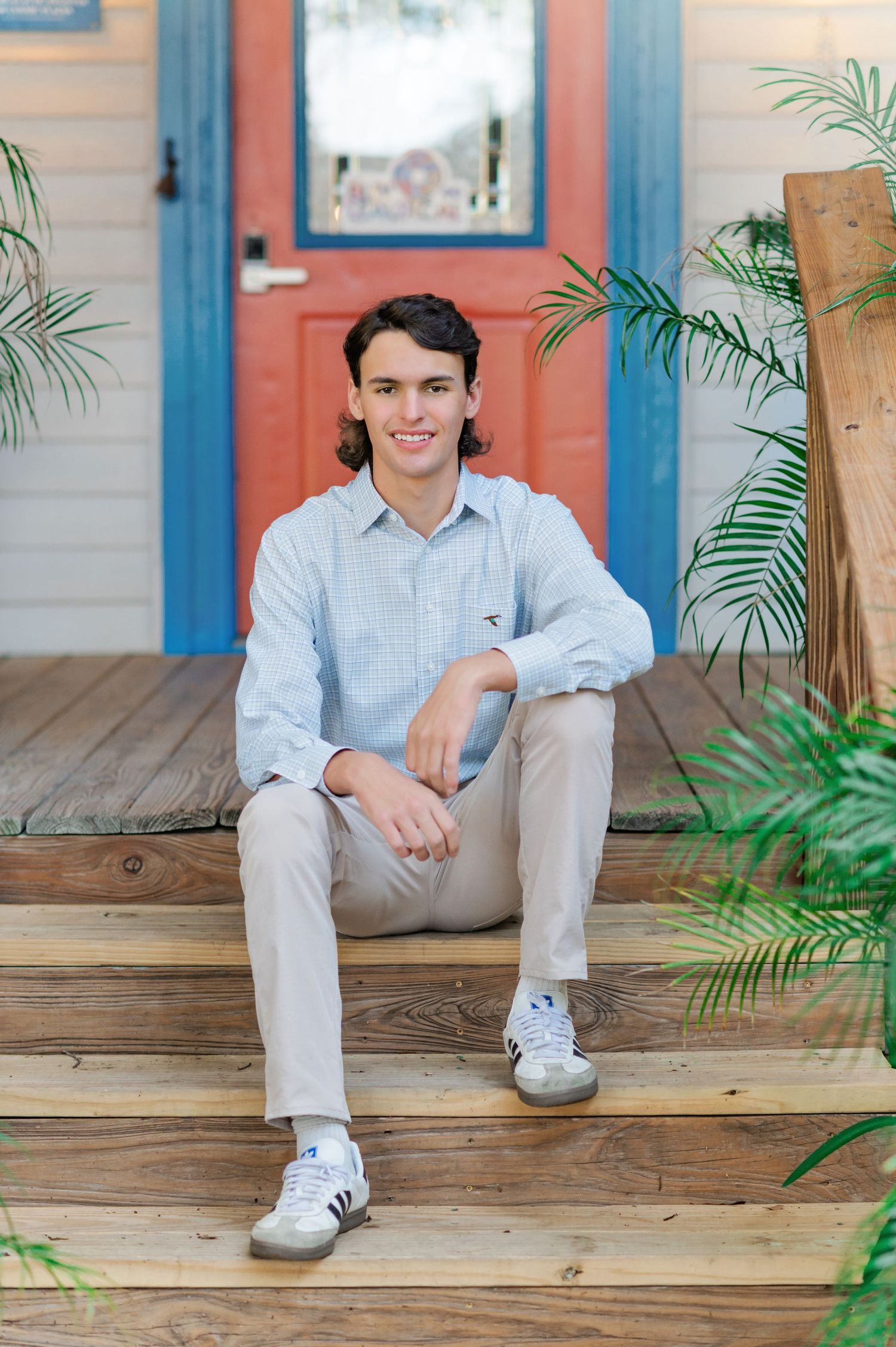 Young man in light blue shirt and khakis sits smiling on wooden porch steps with tropical plants and red door.