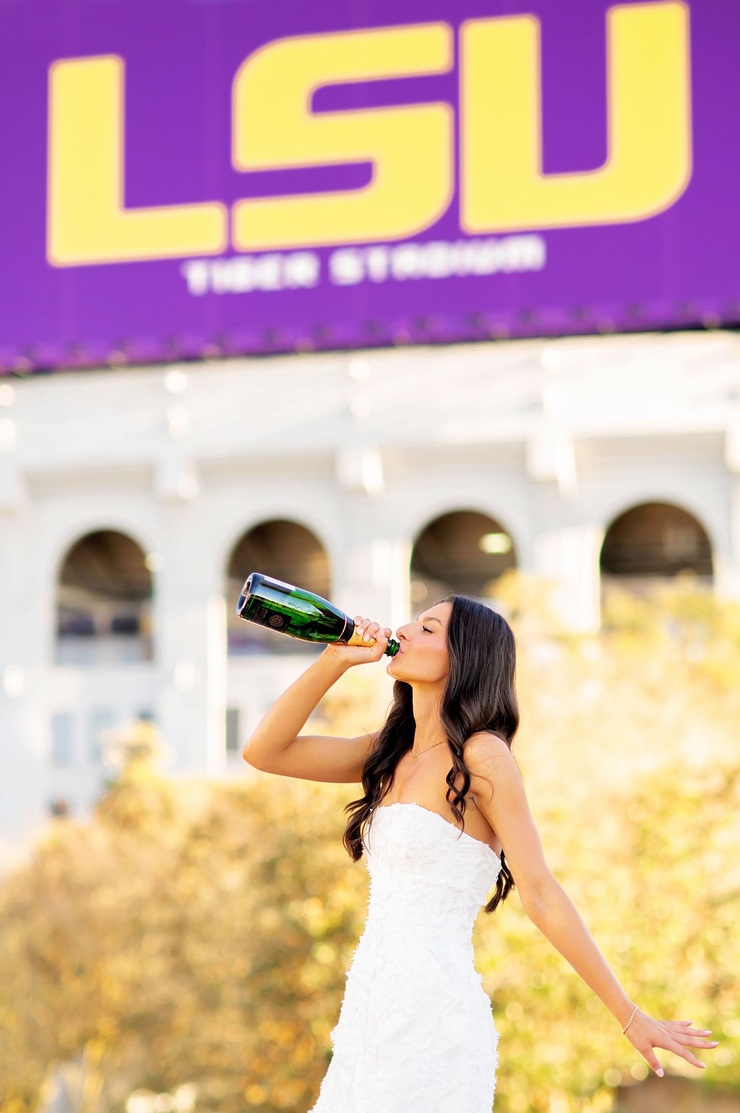 LSU graduate in white dress celebrating with champagne bottle in front of Tiger Stadium sign.