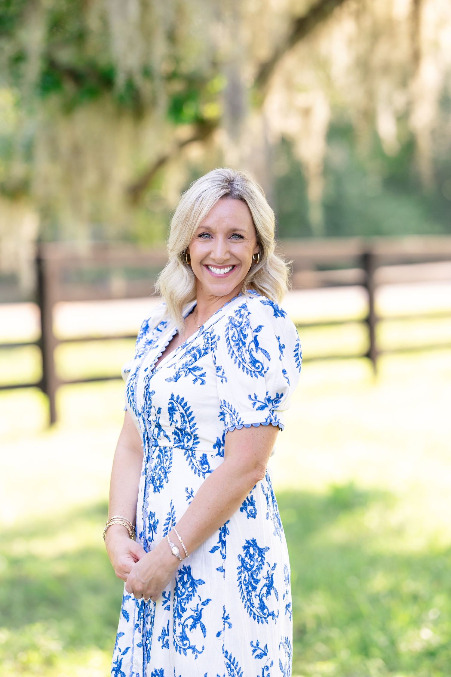 Cheerful blonde woman in blue floral dress smiles outdoors near fence with Spanish moss trees.