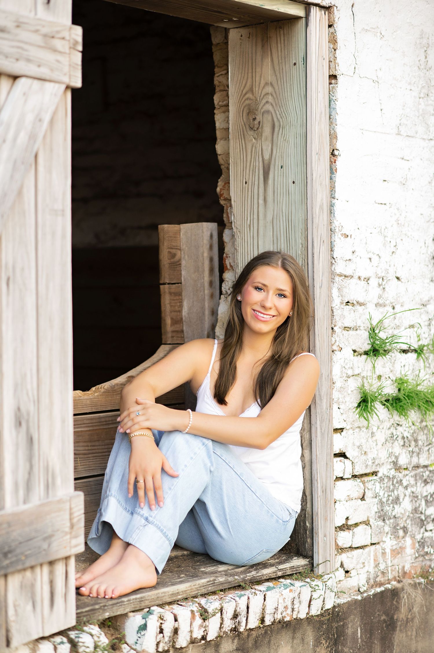 Smiling young woman in white tank and jeans sitting barefoot in rustic wooden doorway of old building.