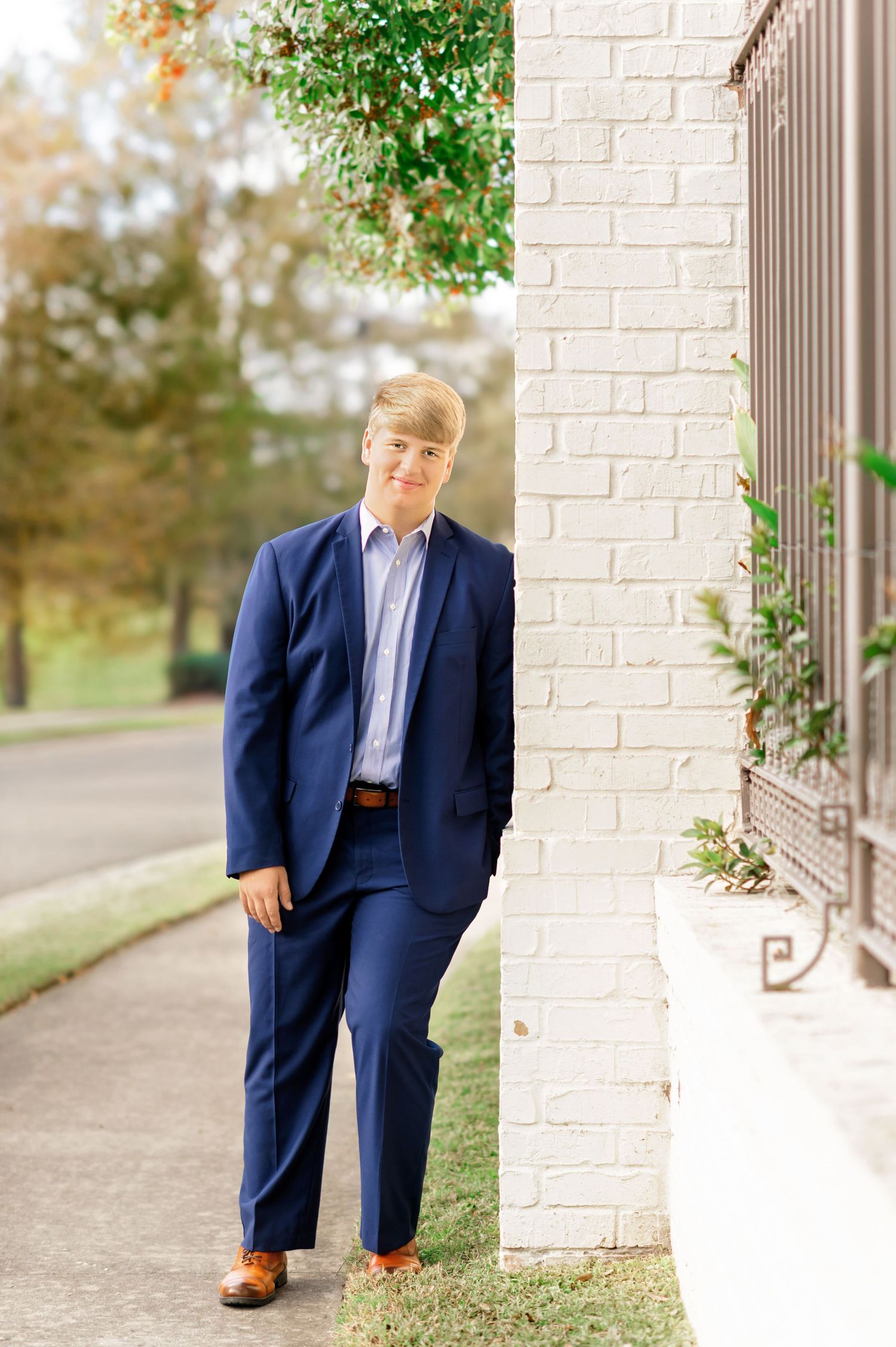 Teen boy in navy suit leans against white brick wall on tree-lined sidewalk in classic senior portrait.