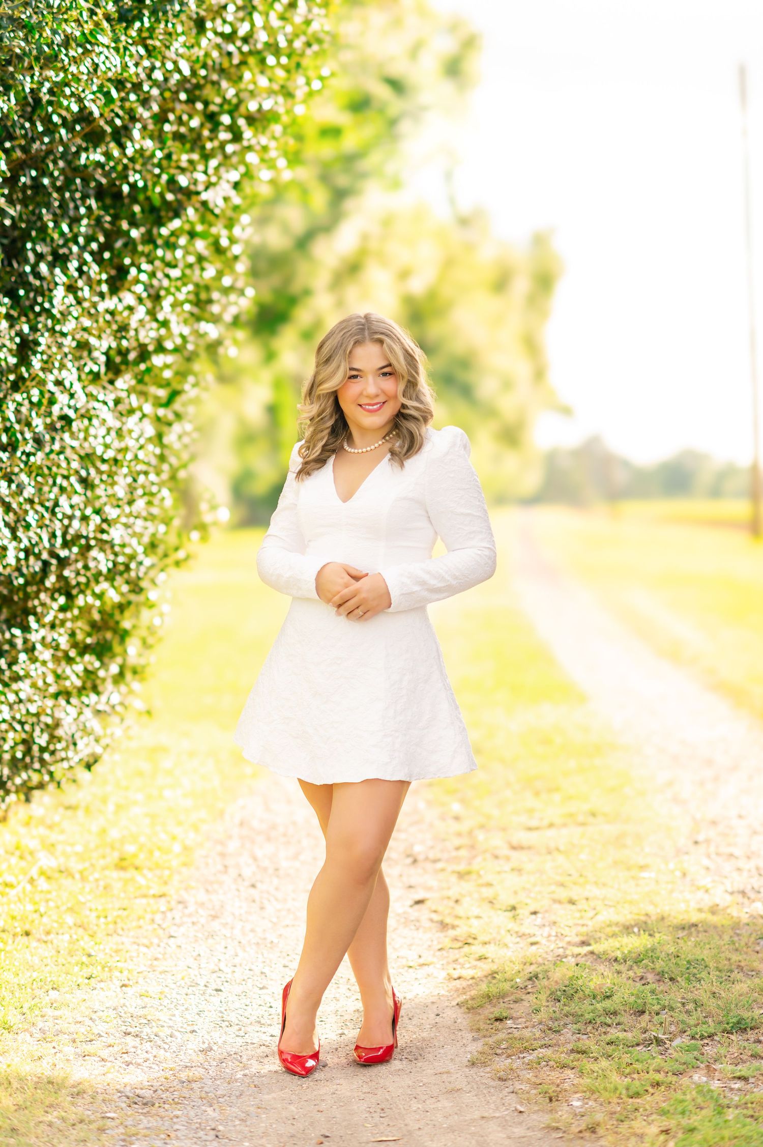Blonde teen girl in white long-sleeve mini dress with red heels posing on a sunny garden path.