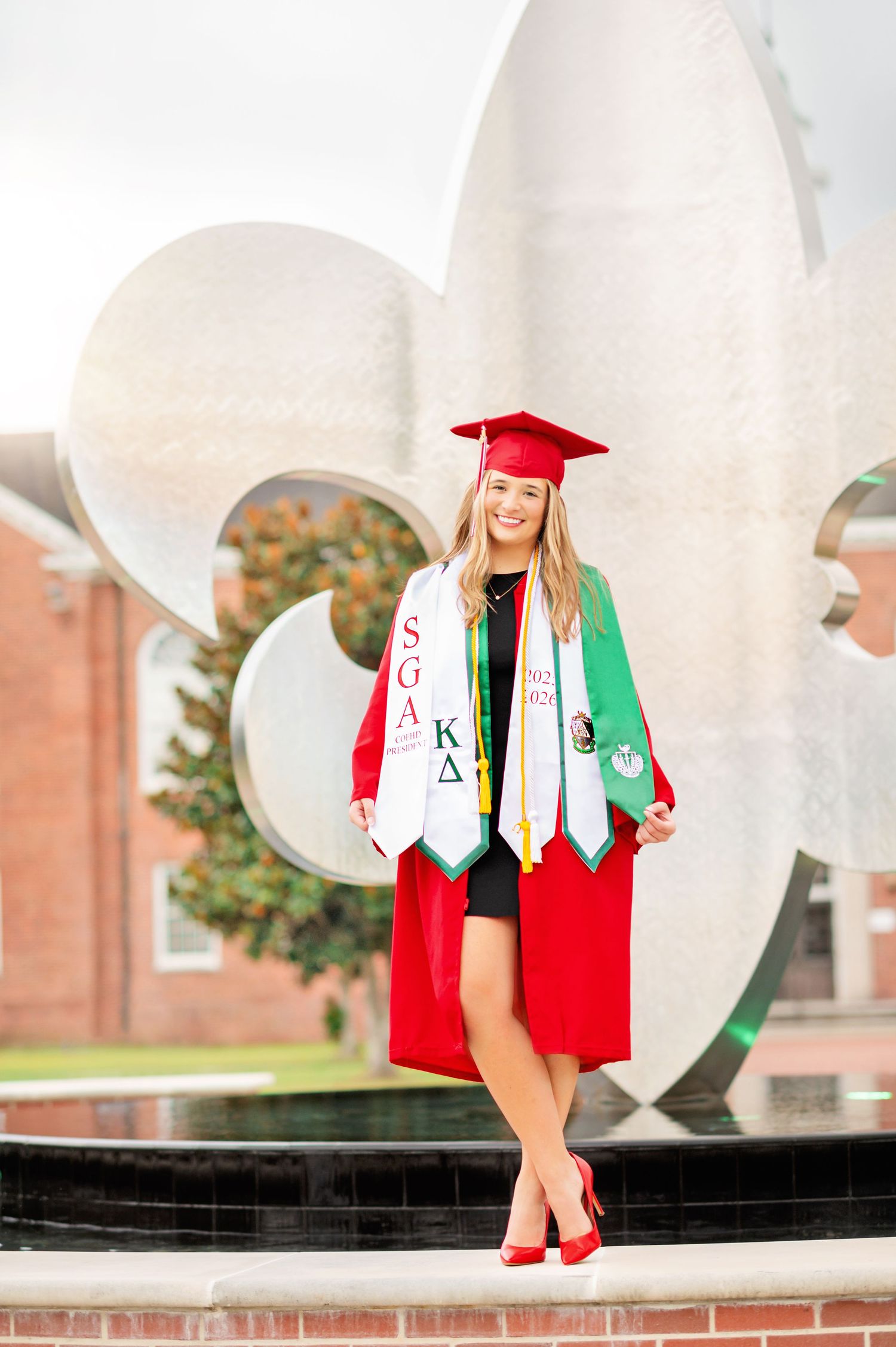 Smiling female graduate in red cap and gown with SGA and Kappa Delta stoles poses outdoors at university campus.