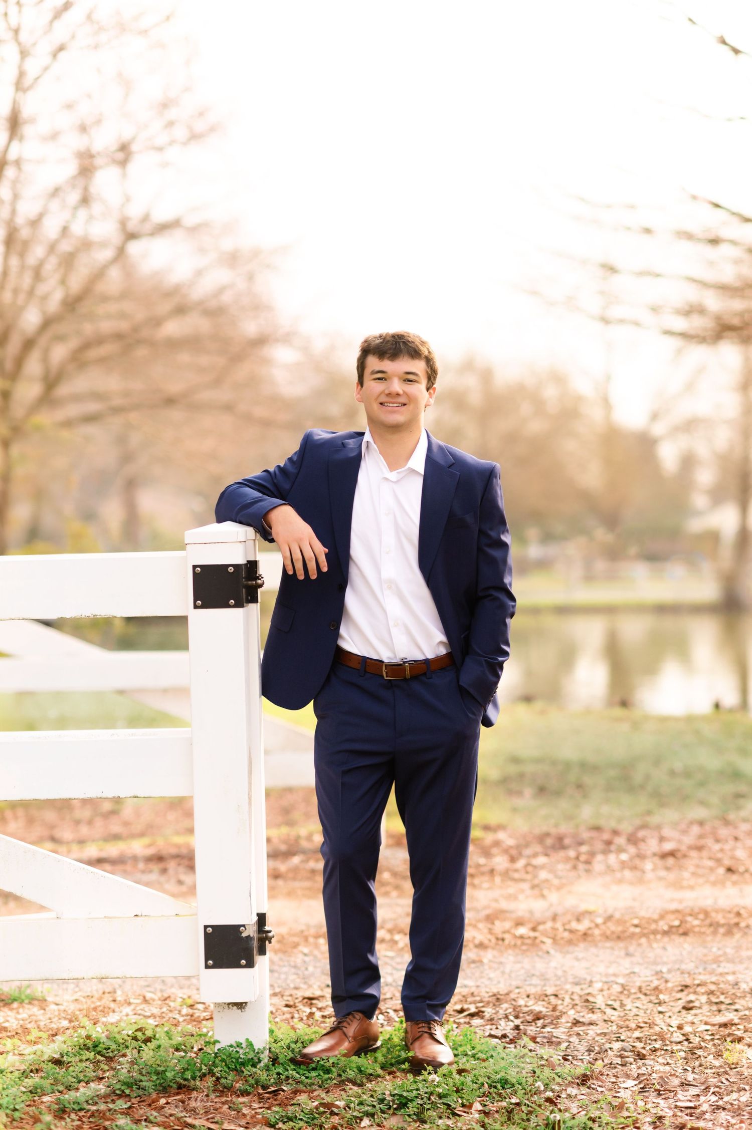 Young man in navy suit leaning on white fence outdoors, smiling confidently in a park setting.