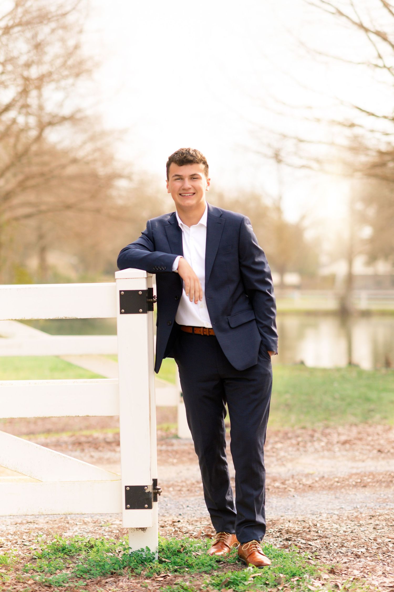 Young man in navy suit leaning on white fence, smiling outdoors in a park setting with trees and pond.