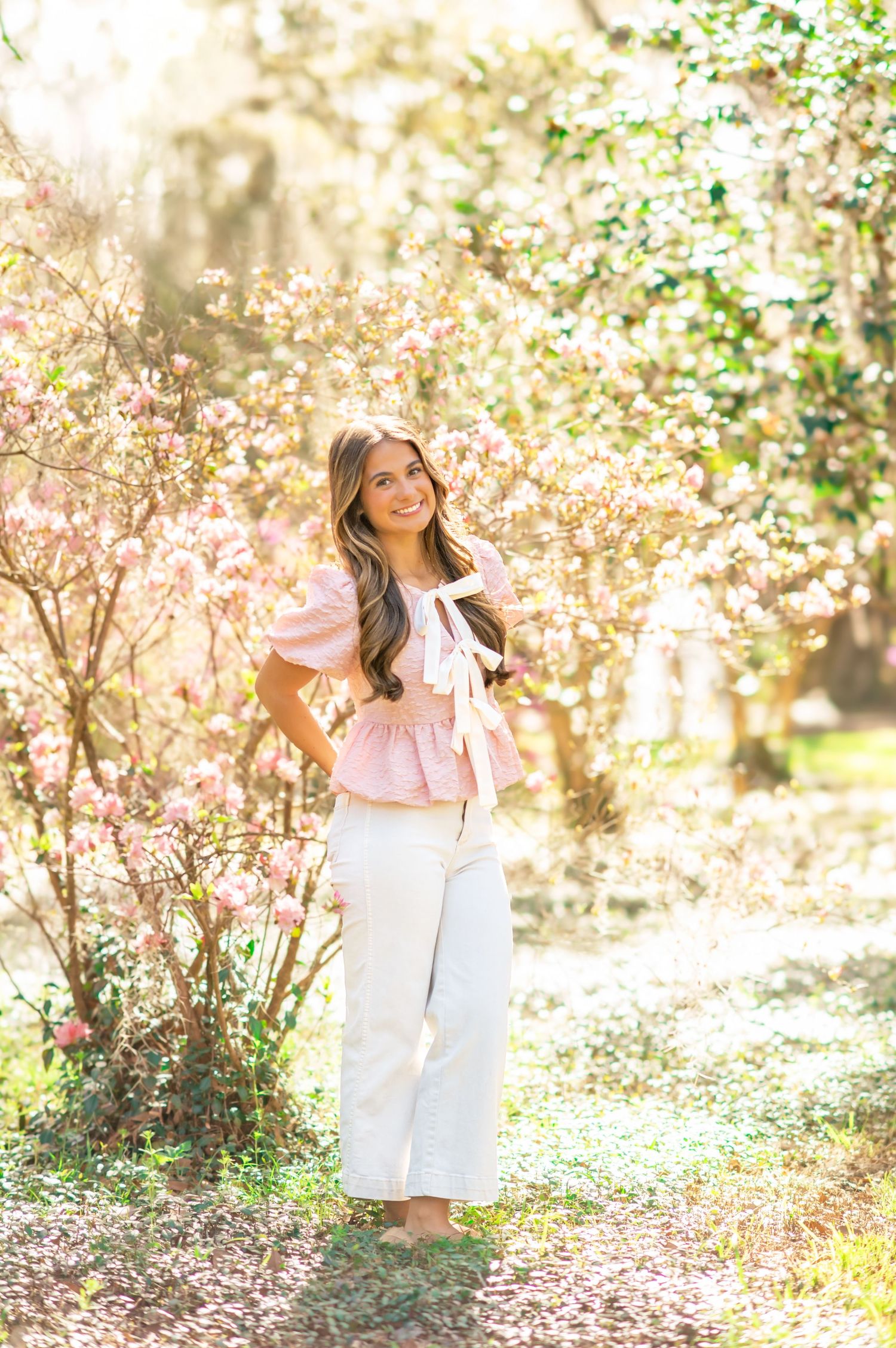 Brunette woman in pink top and white pants stands among pink magnolia blossoms in golden sunlight.
