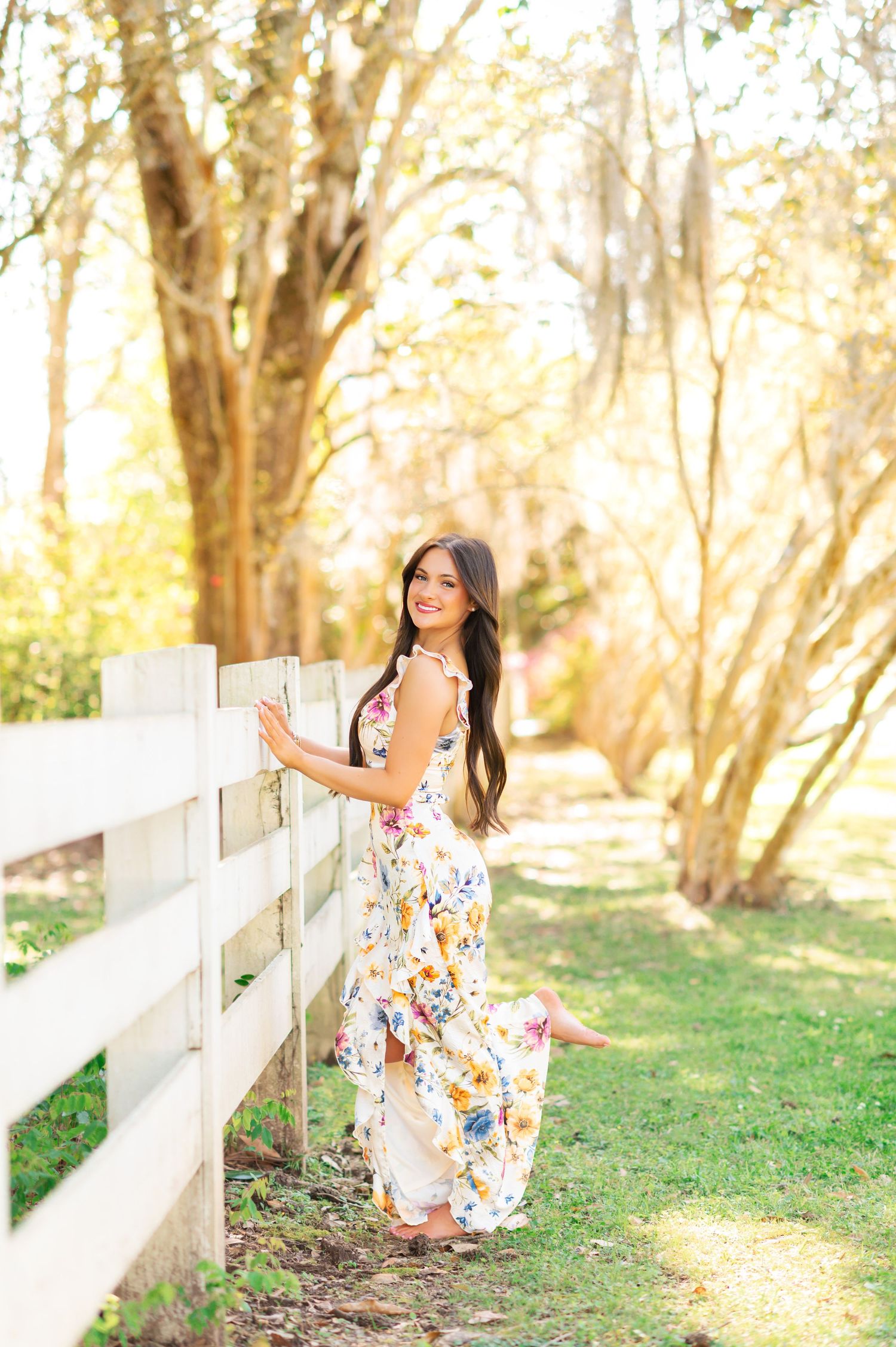 Smiling brunette woman in floral maxi dress leans against white wooden fence in sunny garden.