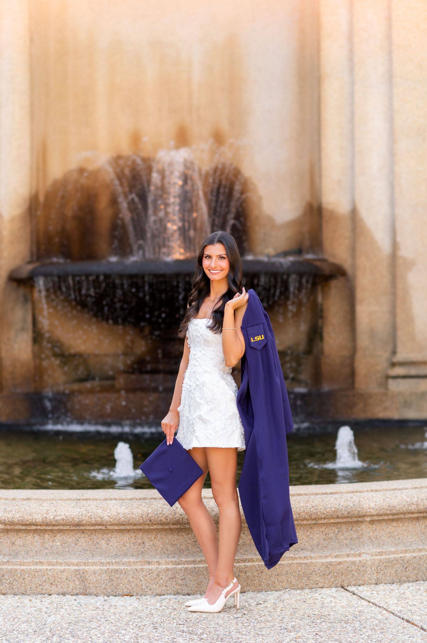 Young woman in white dress holding purple graduation gown and cap in front of a fountain.