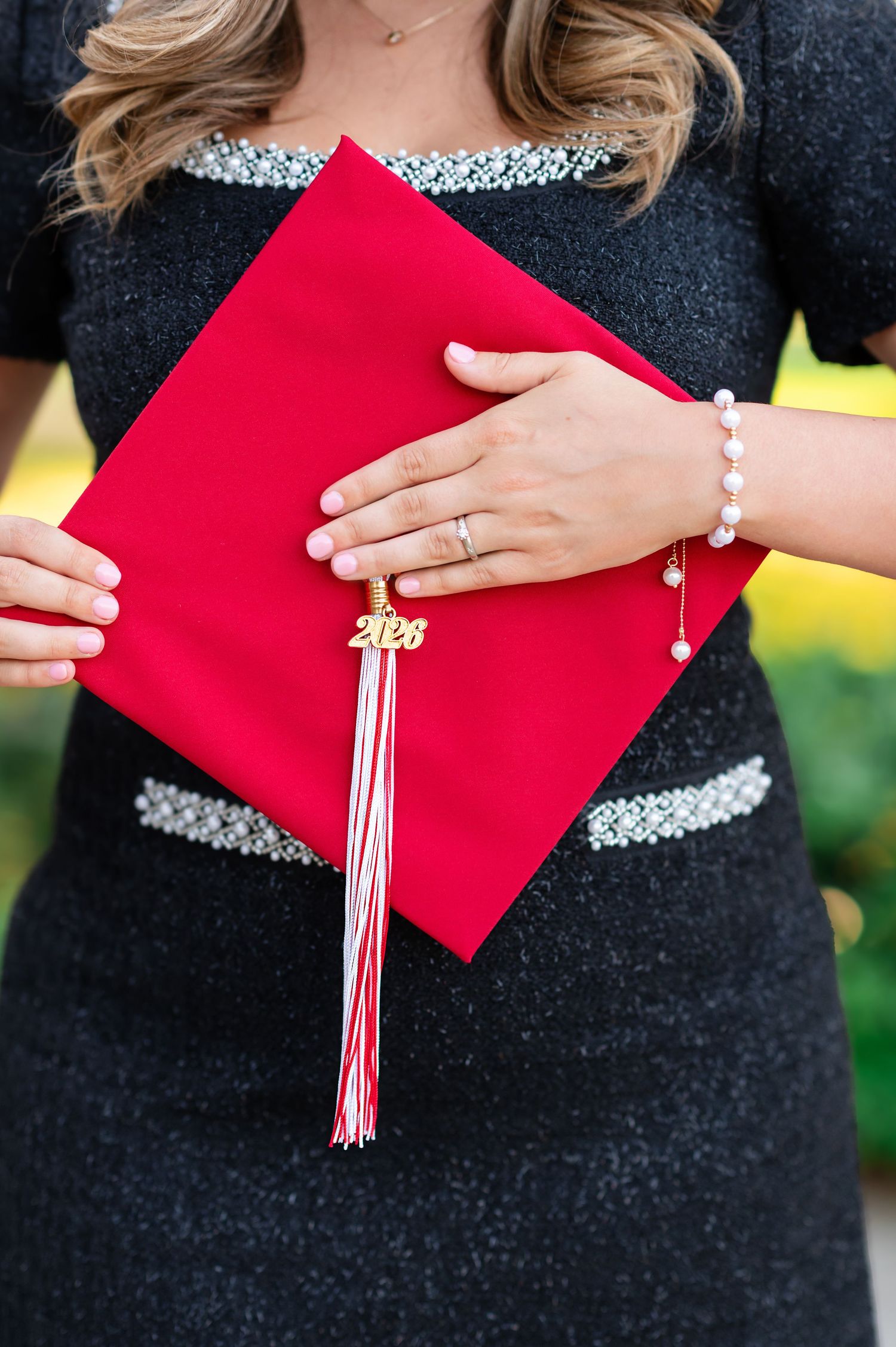 Close-up of a graduate holding a red mortarboard cap with a gold 2026 tassel, wearing a pearl bracelet and ring.
