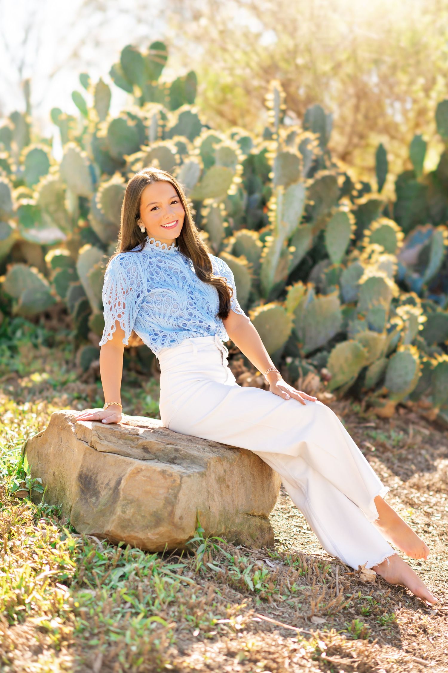 Smiling young woman in blue lace top and white pants sitting on rock near large green cactus plants.