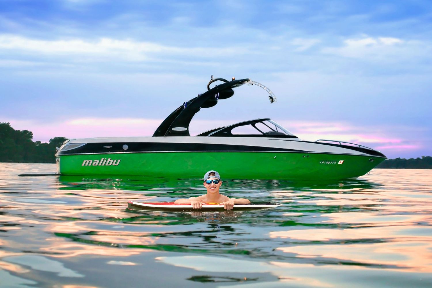 Person swimming near a green Malibu wakeboard boat on calm lake water at sunset.
