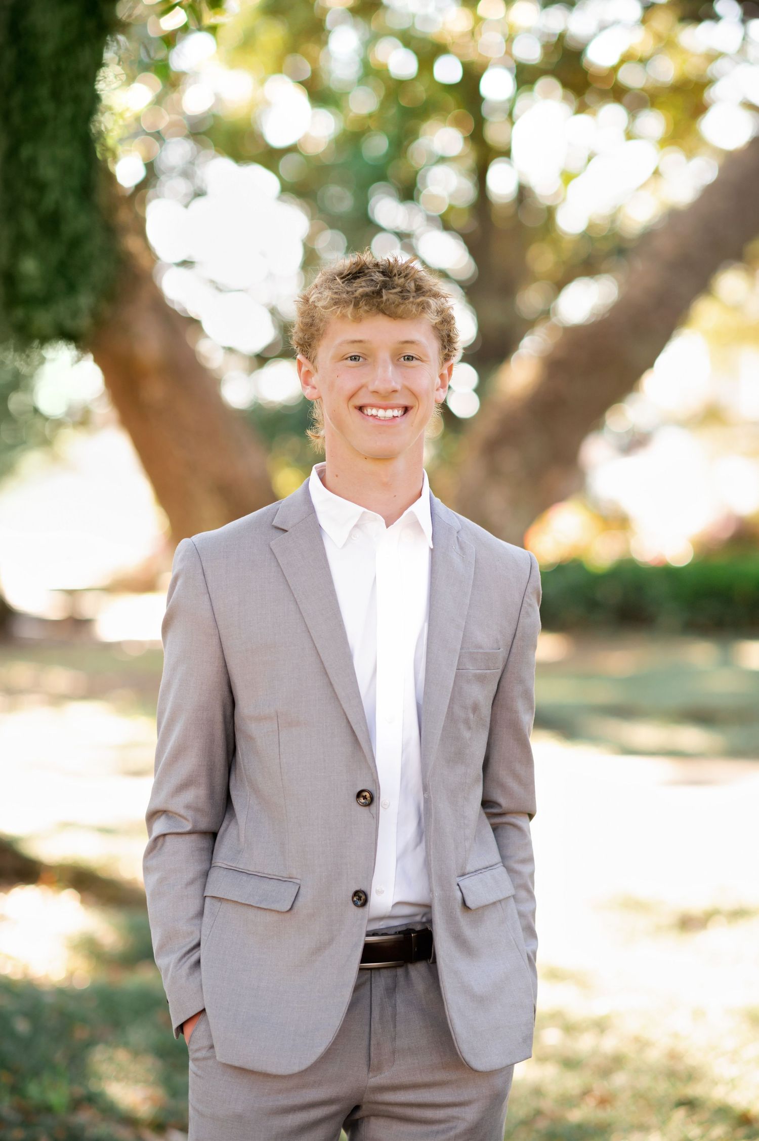 Young man in light gray suit smiling outdoors in a sunny park setting for a formal portrait photo.