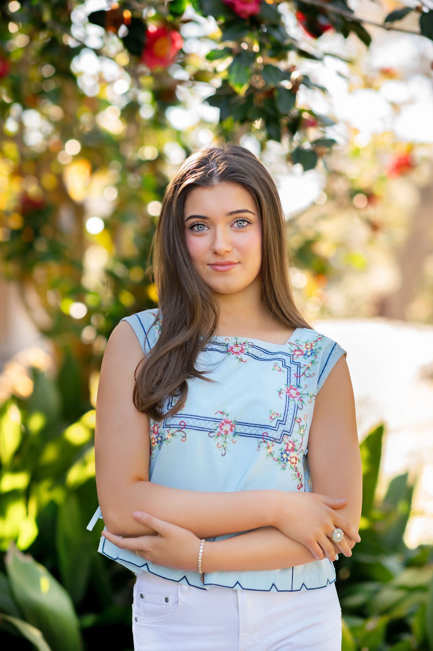 Young woman in light blue floral top with arms crossed, smiling outdoors among green foliage and flowers.