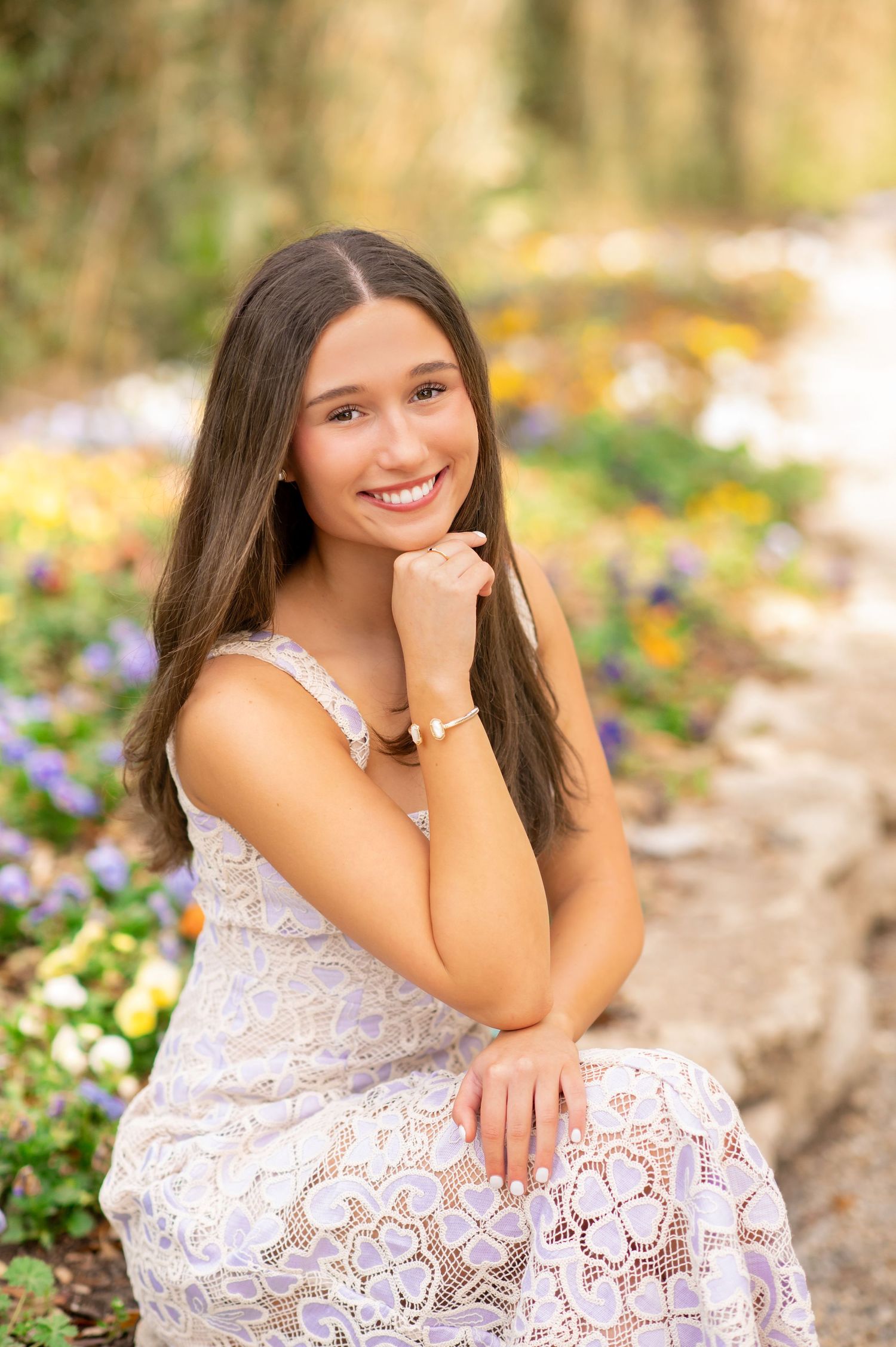 Young woman in white lace dress smiling in a colorful flower garden during a senior portrait session.