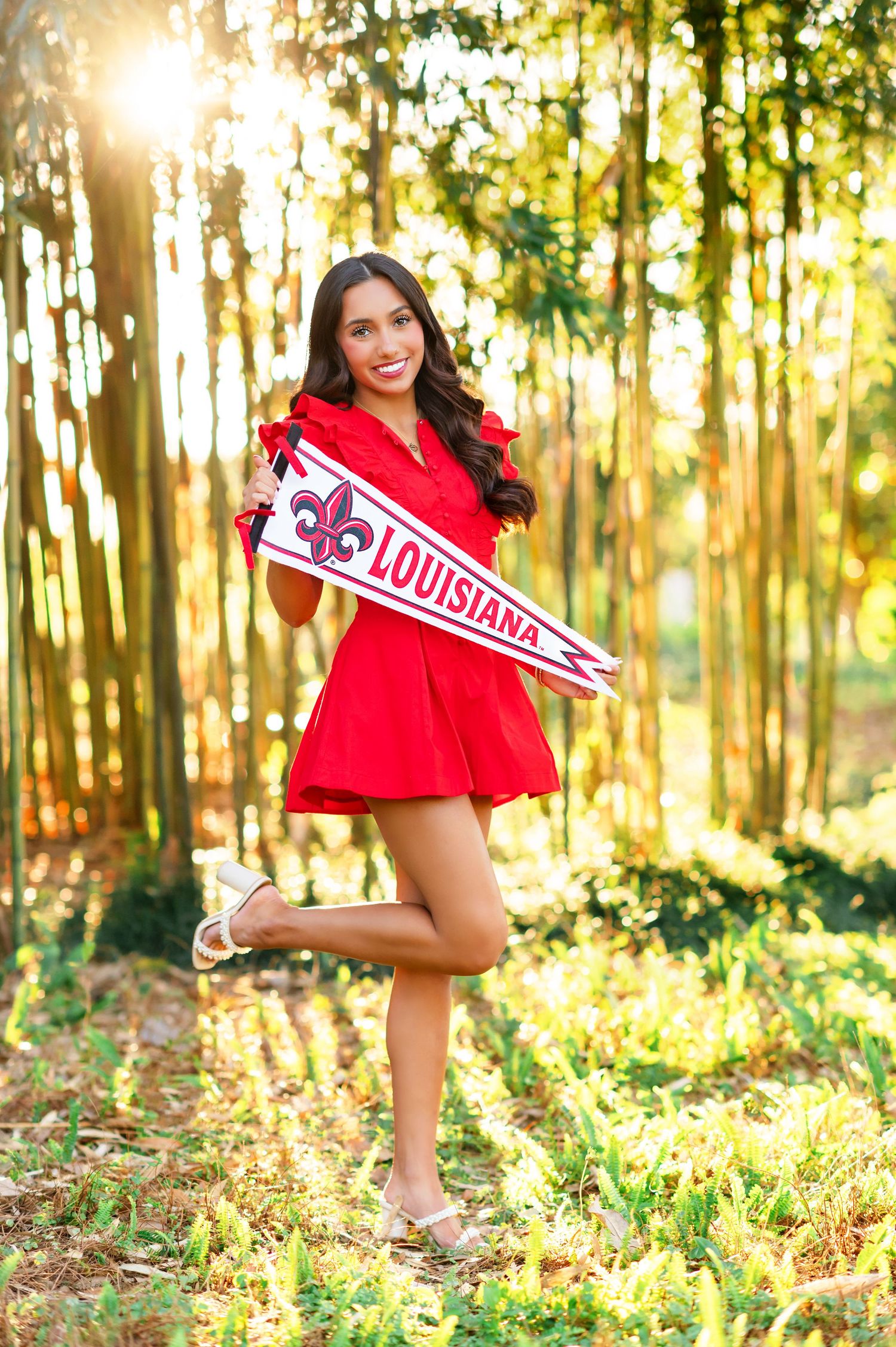 A smiling young woman in a red dress holds a Louisiana Ragin Cajuns pennant flag outdoors in a sunlit forest setting.