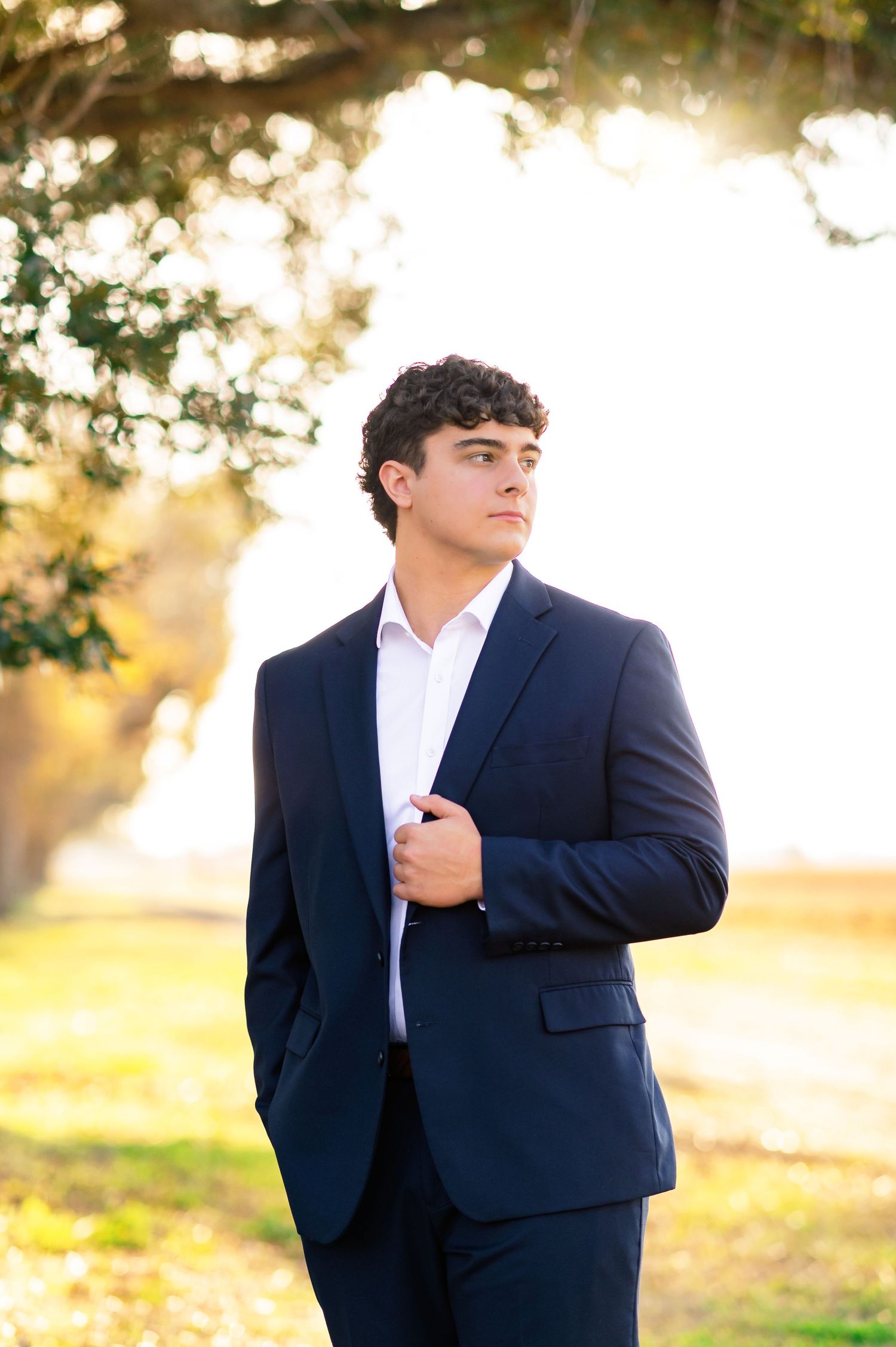 Young man in navy blue suit posing outdoors under a tree with golden sunlight in background.