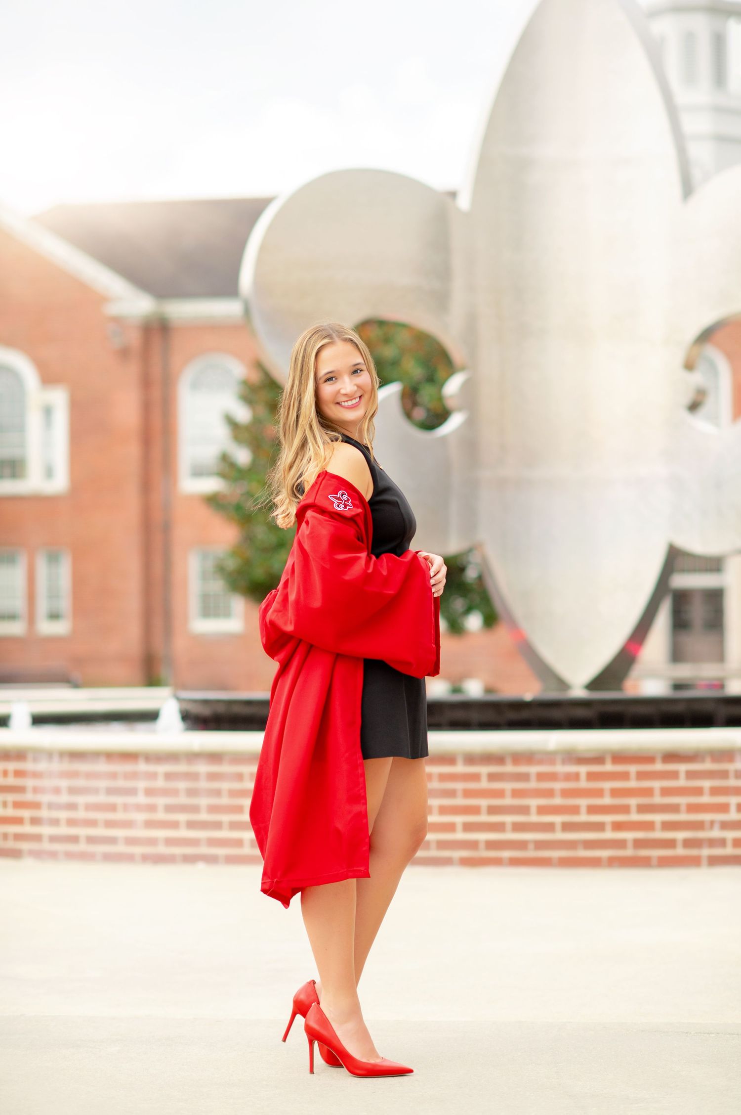 Young blonde woman in red graduation gown and heels smiles outdoors near a brick building and silver sculpture.