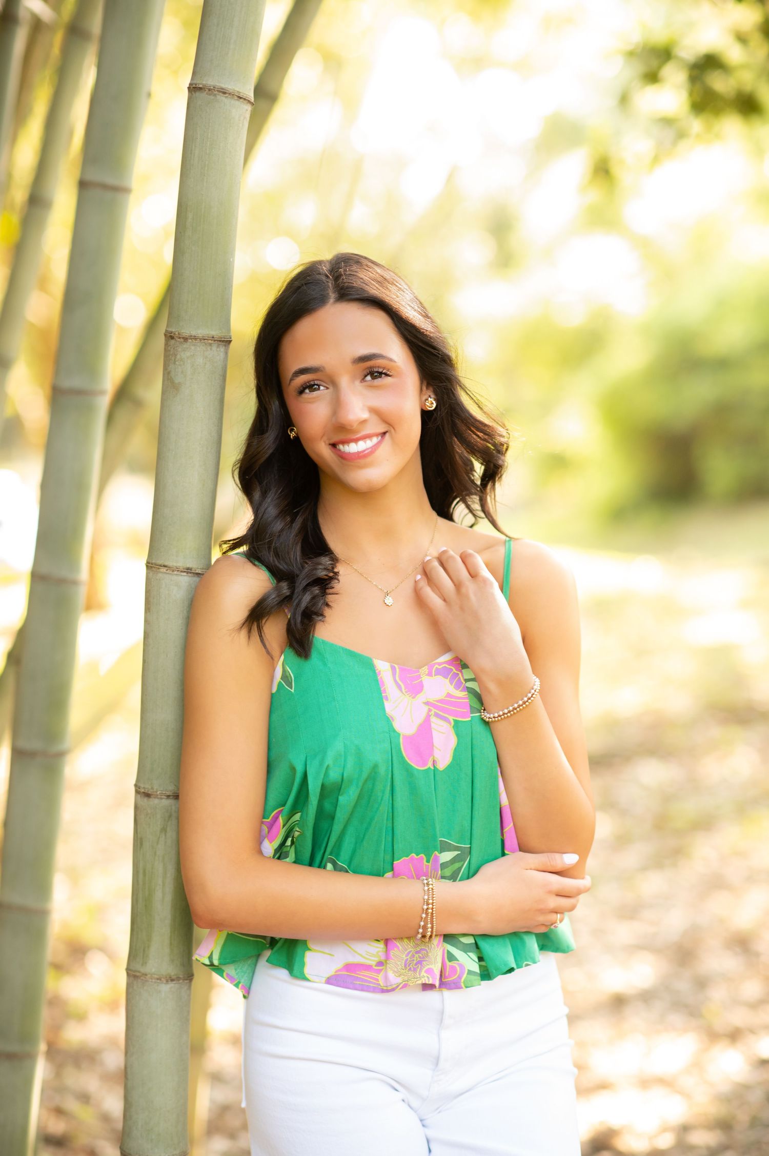 Young woman in green floral top and white pants smiling beside bamboo trees in a sunlit outdoor setting.