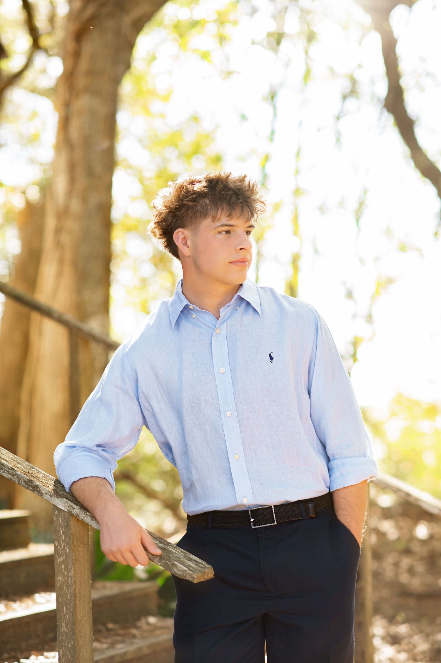 Young man in light blue Ralph Lauren shirt and dark pants leaning on wooden railing outdoors in sunlit park.