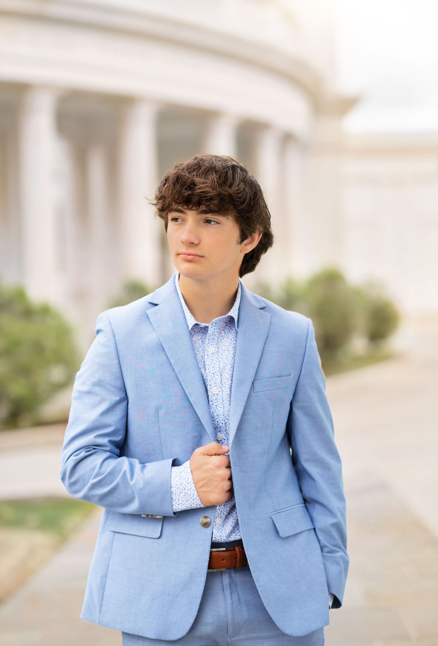 Young man in light blue suit poses outdoors near classical columns for a formal portrait photo.
