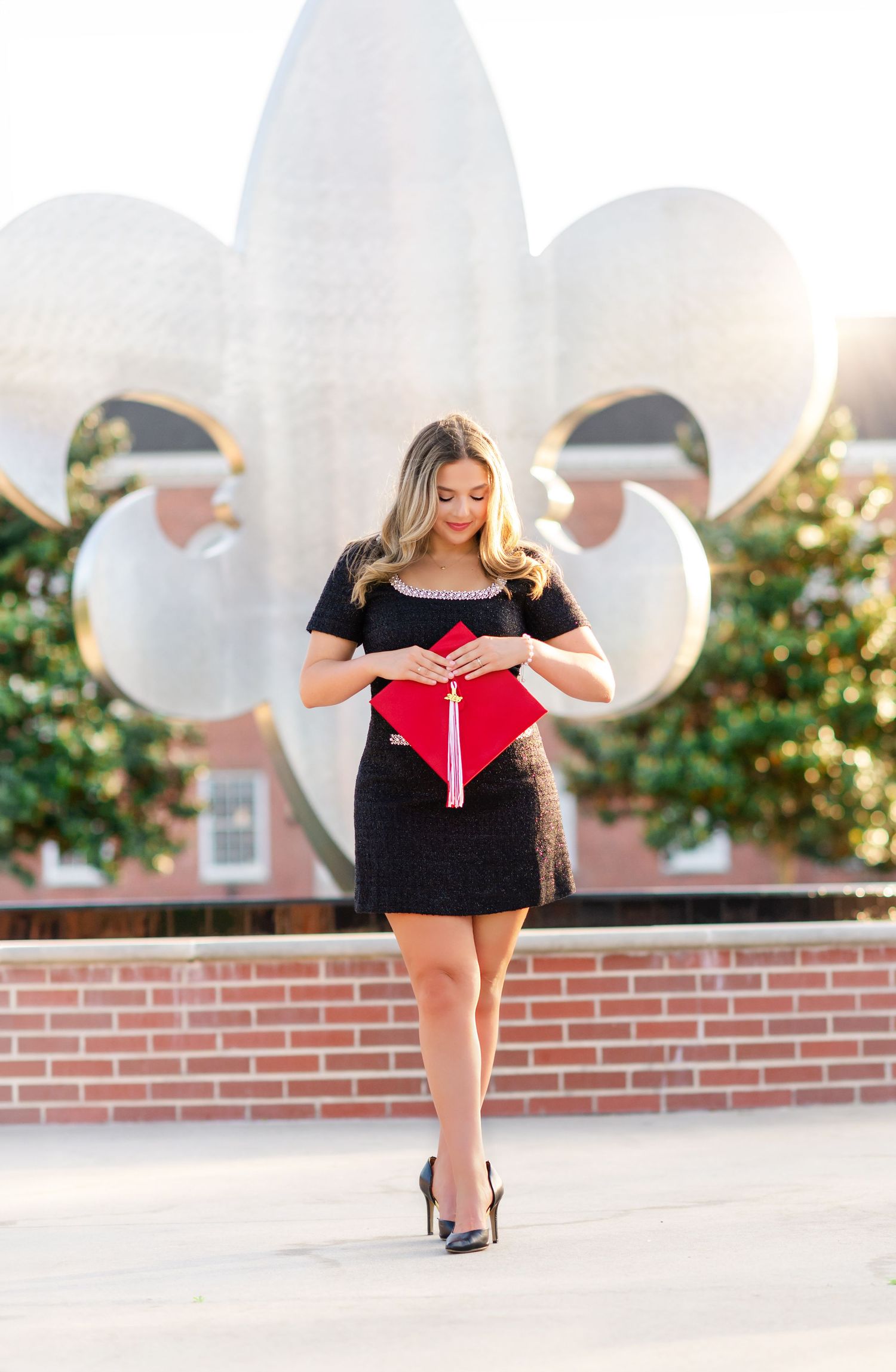 Graduate in black dress holding red graduation cap in front of a fleur-de-lis sculpture outdoors.
