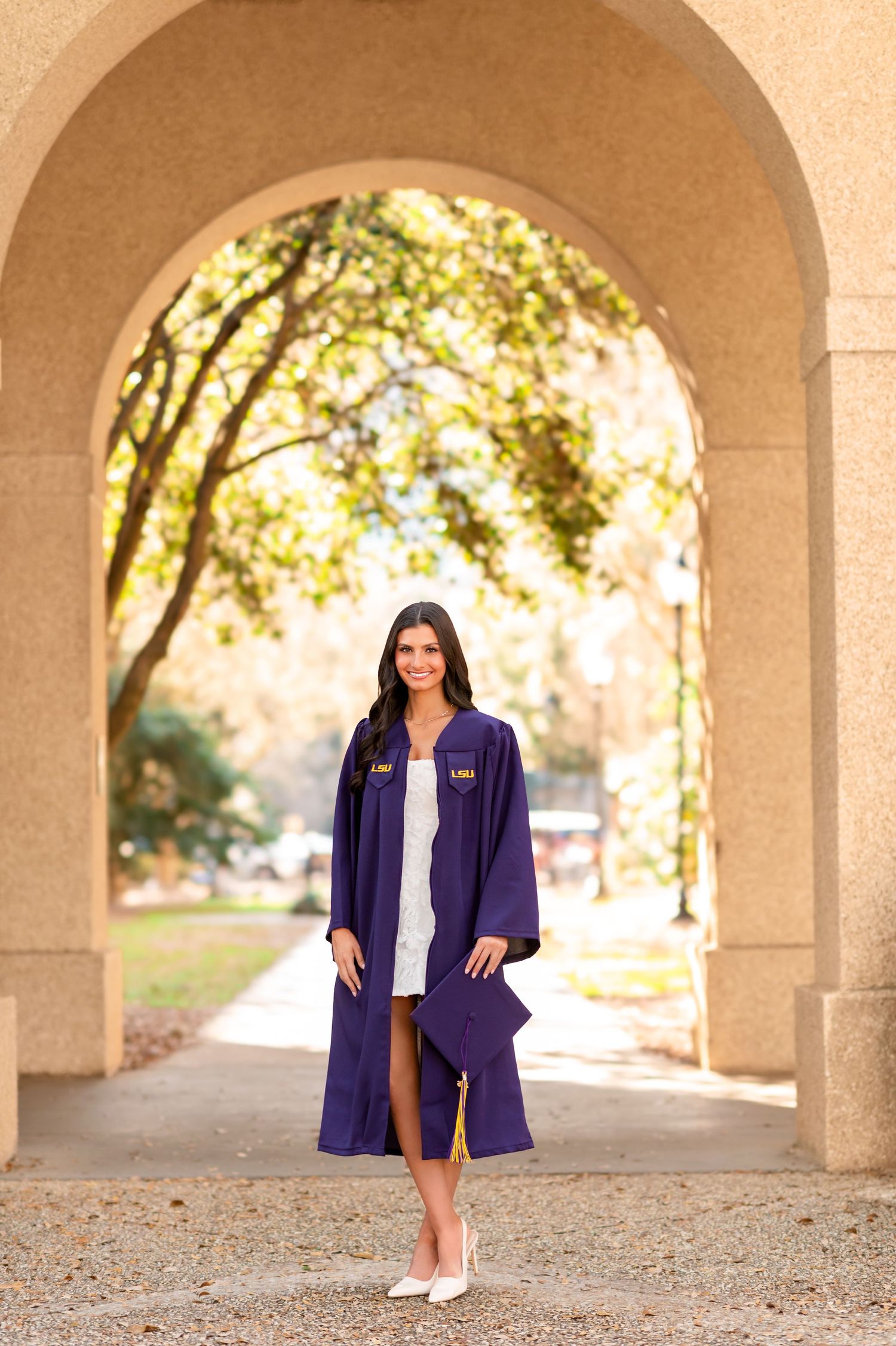 LSU graduate in purple cap and gown poses under stone arch on campus.