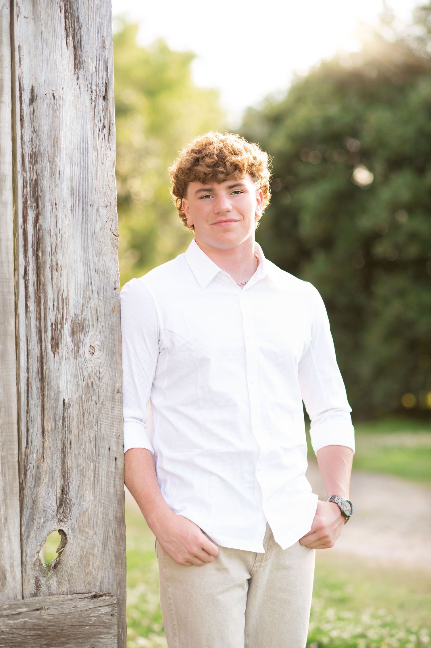 A teenage boy with curly red hair wearing a white shirt and khaki pants leans against a rustic wooden post outdoors.