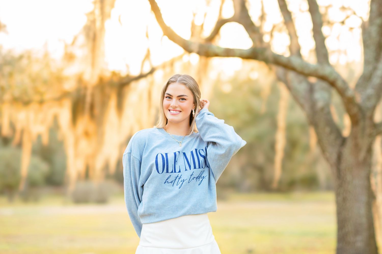 Smiling blonde teen in Ole Miss sweatshirt posing outdoors under Spanish moss-draped trees at sunset.