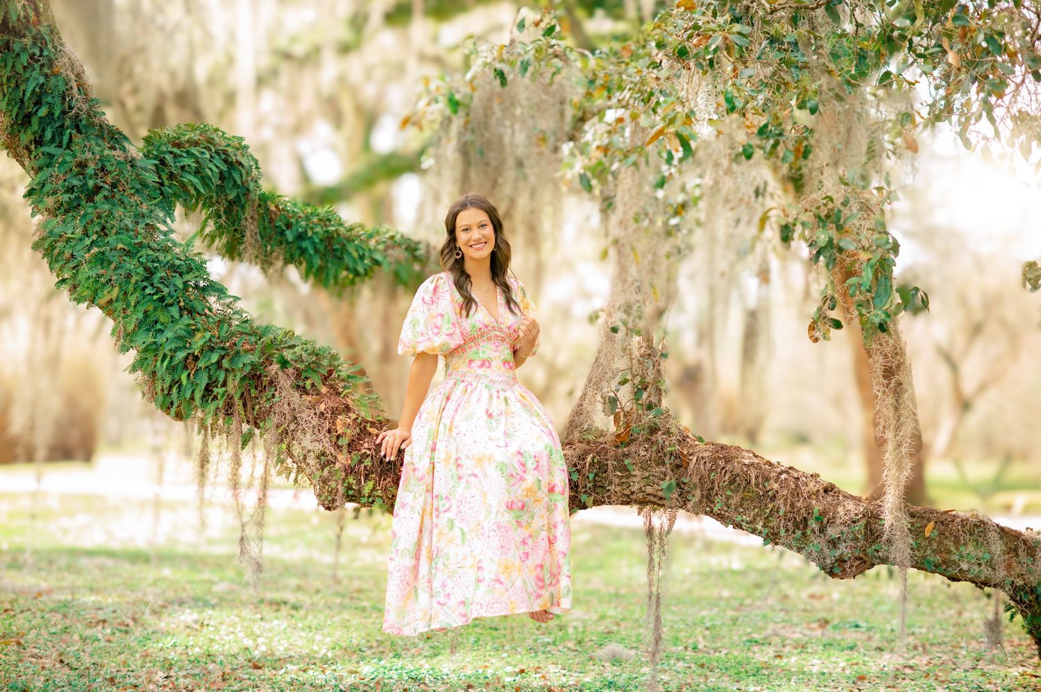 Young woman in floral maxi dress posing on mossy oak tree branch in lush green park setting.