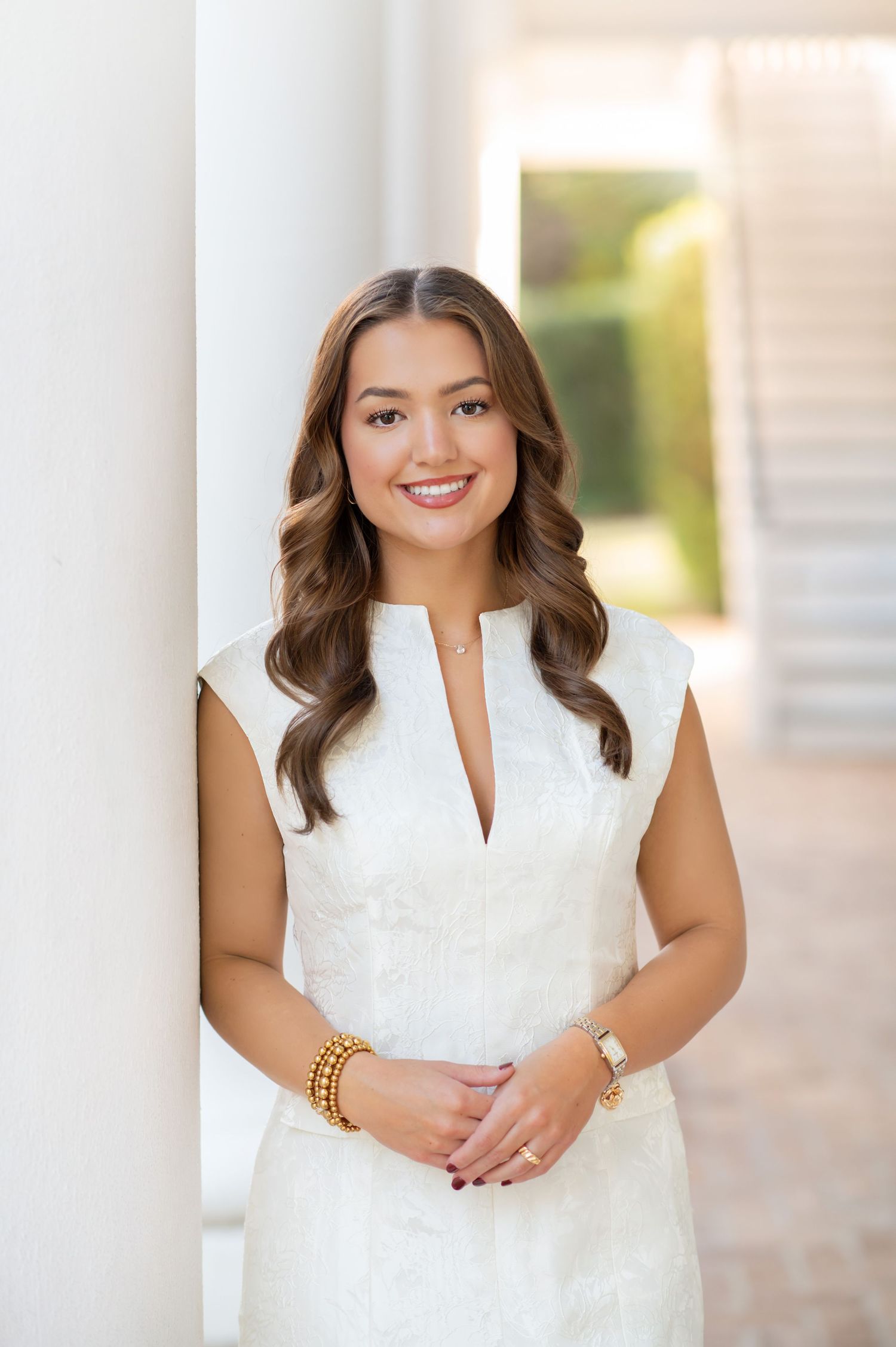 Young woman in white dress leaning against a white column, smiling with wavy brown hair and gold jewelry.