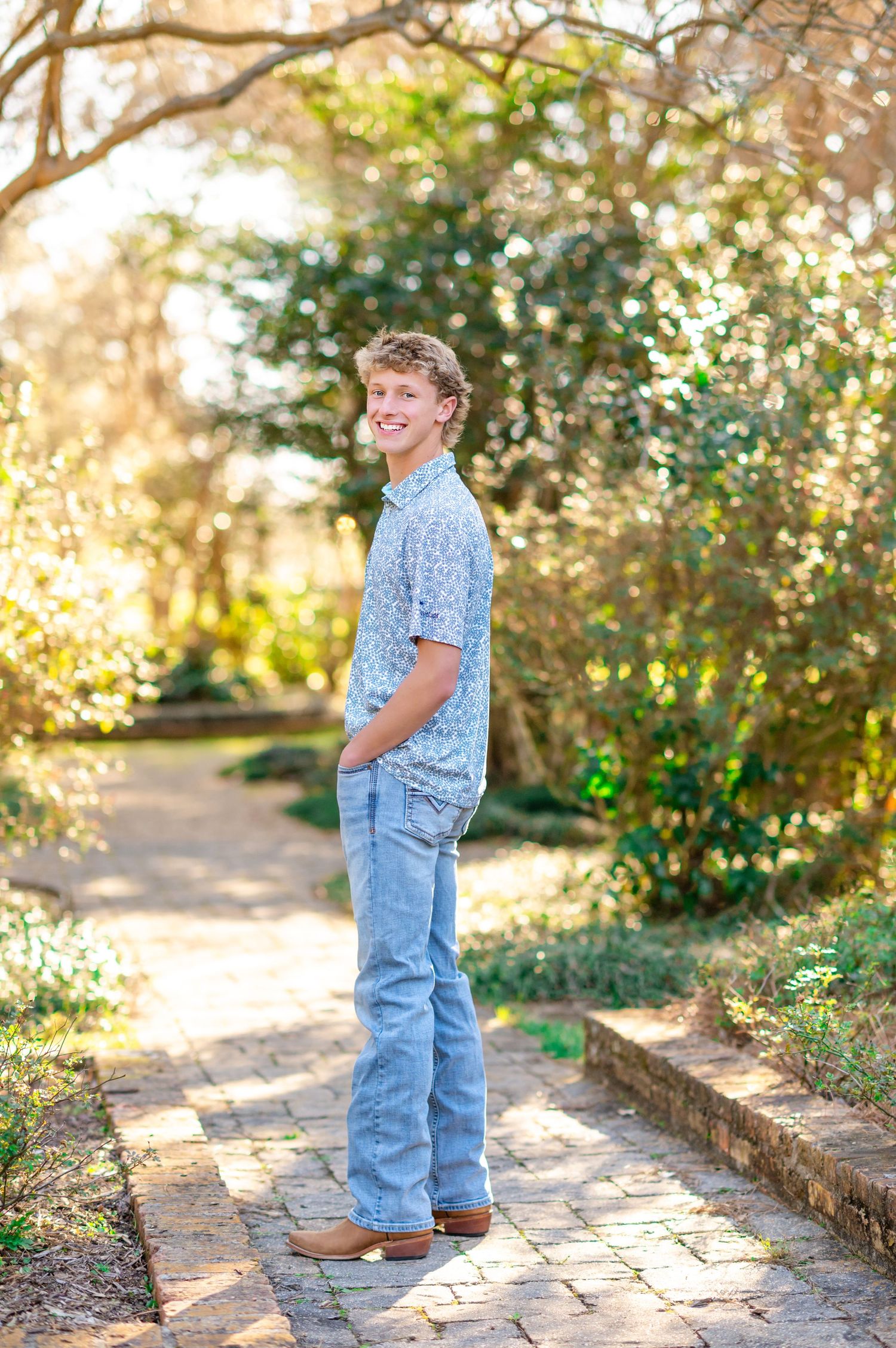 Smiling teenage boy in blue floral shirt and jeans standing on brick path in sunny garden.