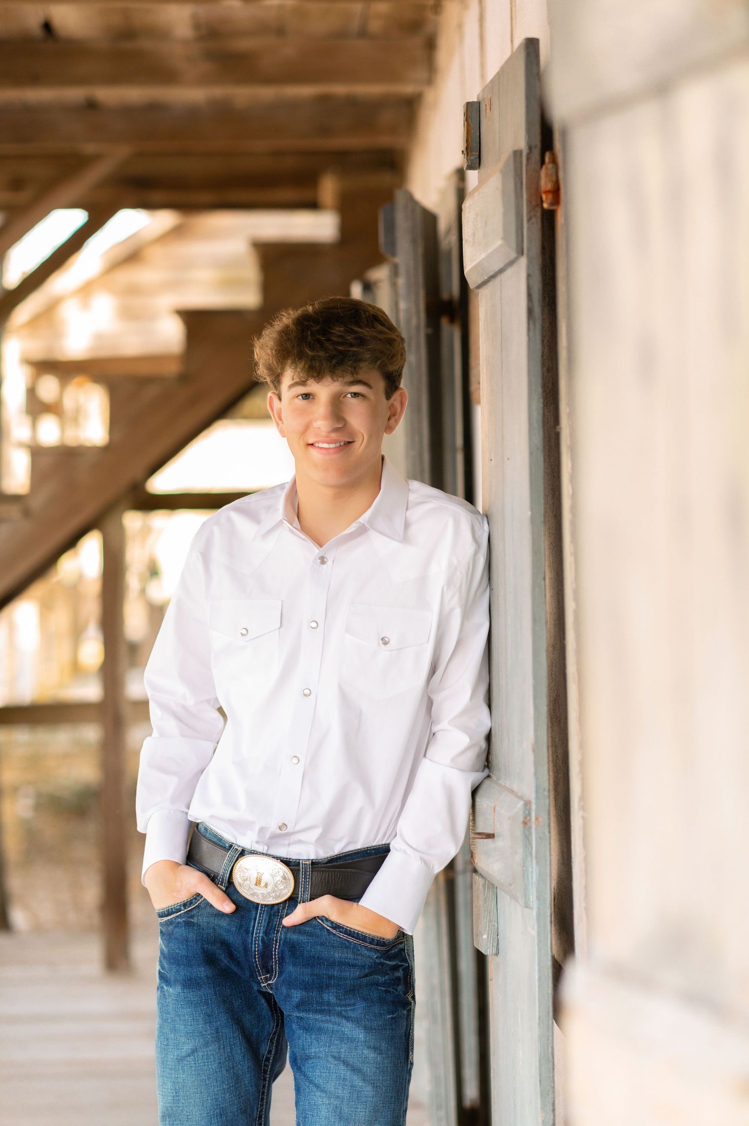 Young man in white western shirt and jeans leaning against barn door smiling.