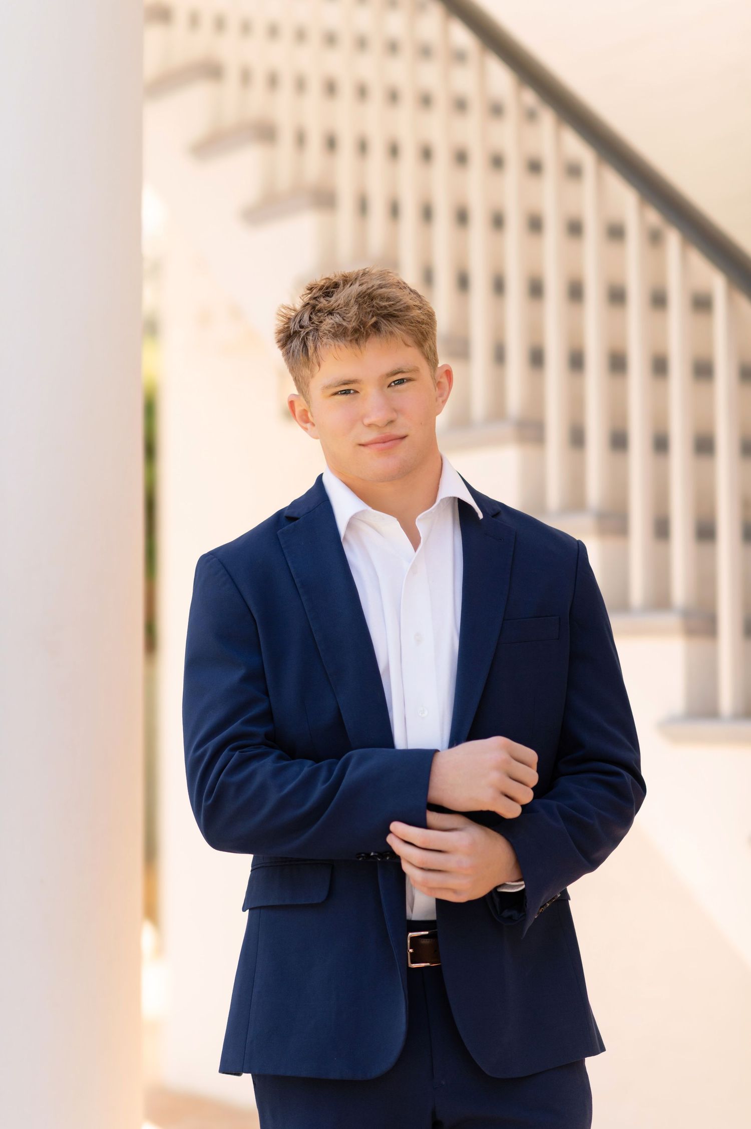Young man in navy blue suit posing confidently near white columns and staircase for senior portrait.